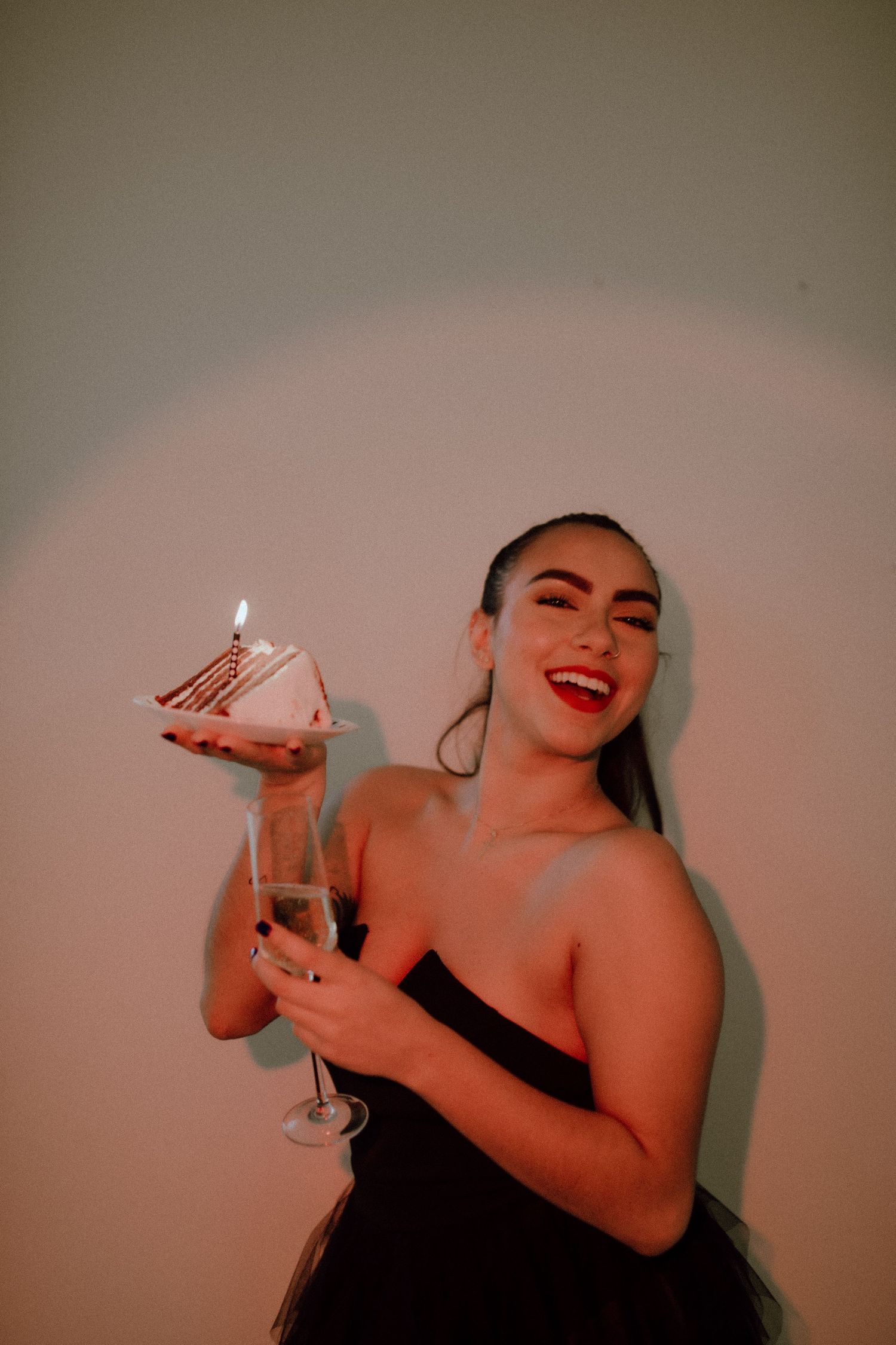 Woman in black dress holds champagne glass and slice of cake with candle.