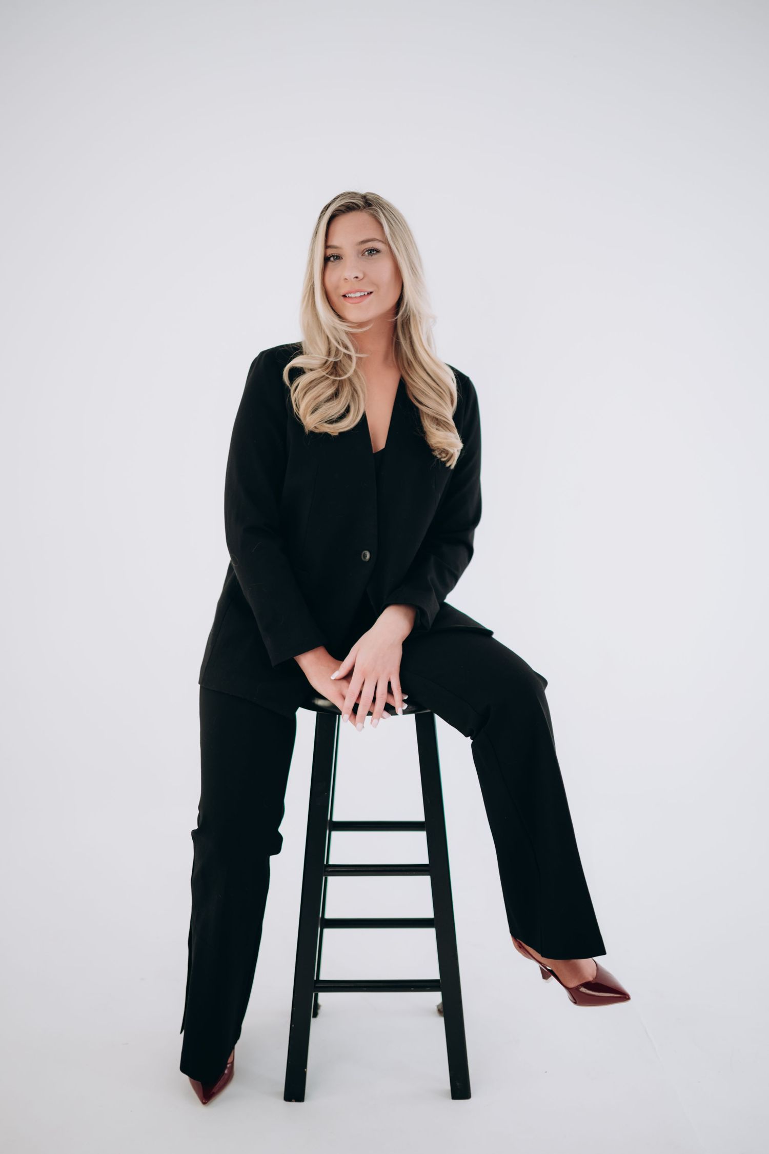 Studio photo in black business suit sitting on wooden stool against white background.