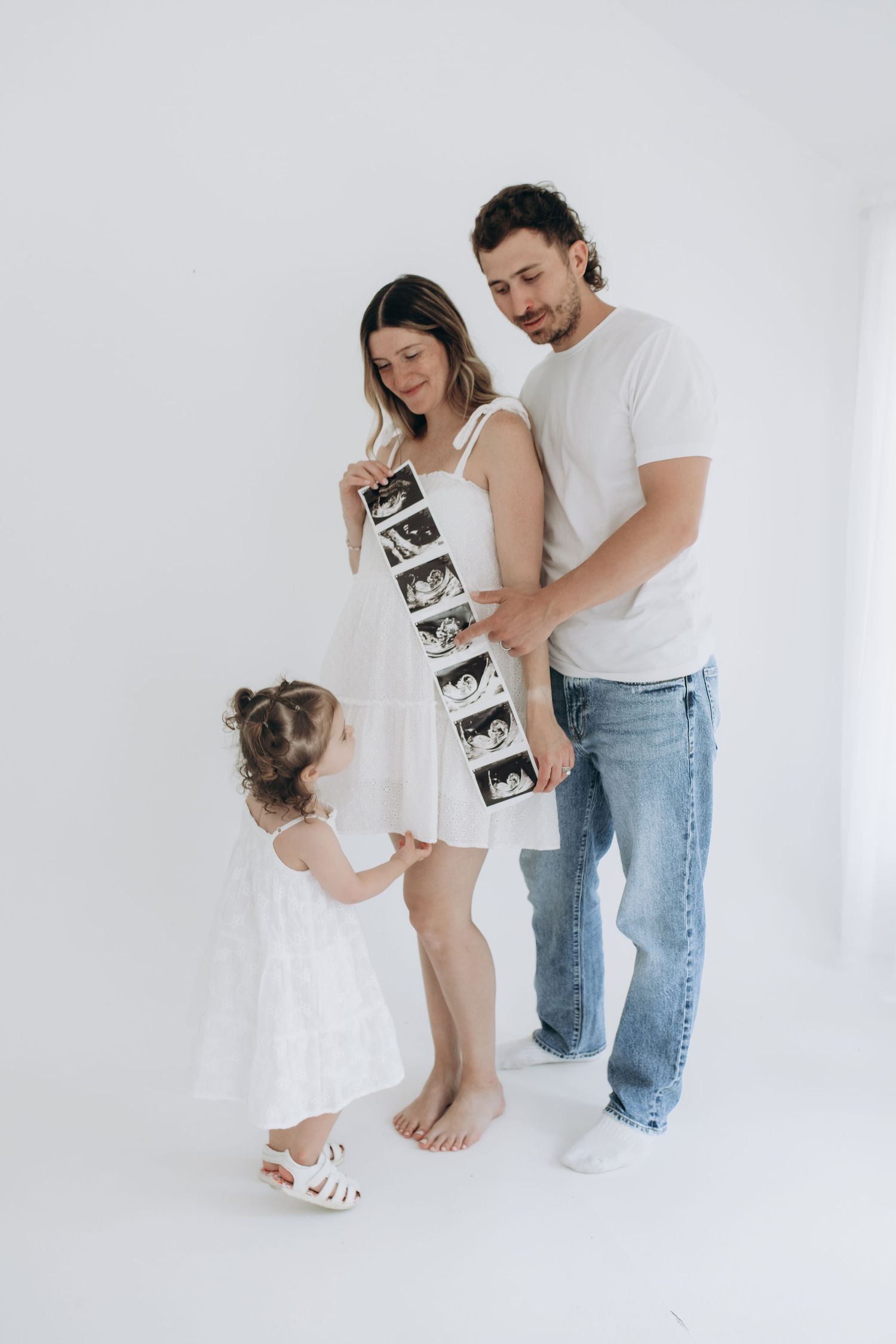 A family holds ultrasound photos while embracing in a minimalist white studio setting.
