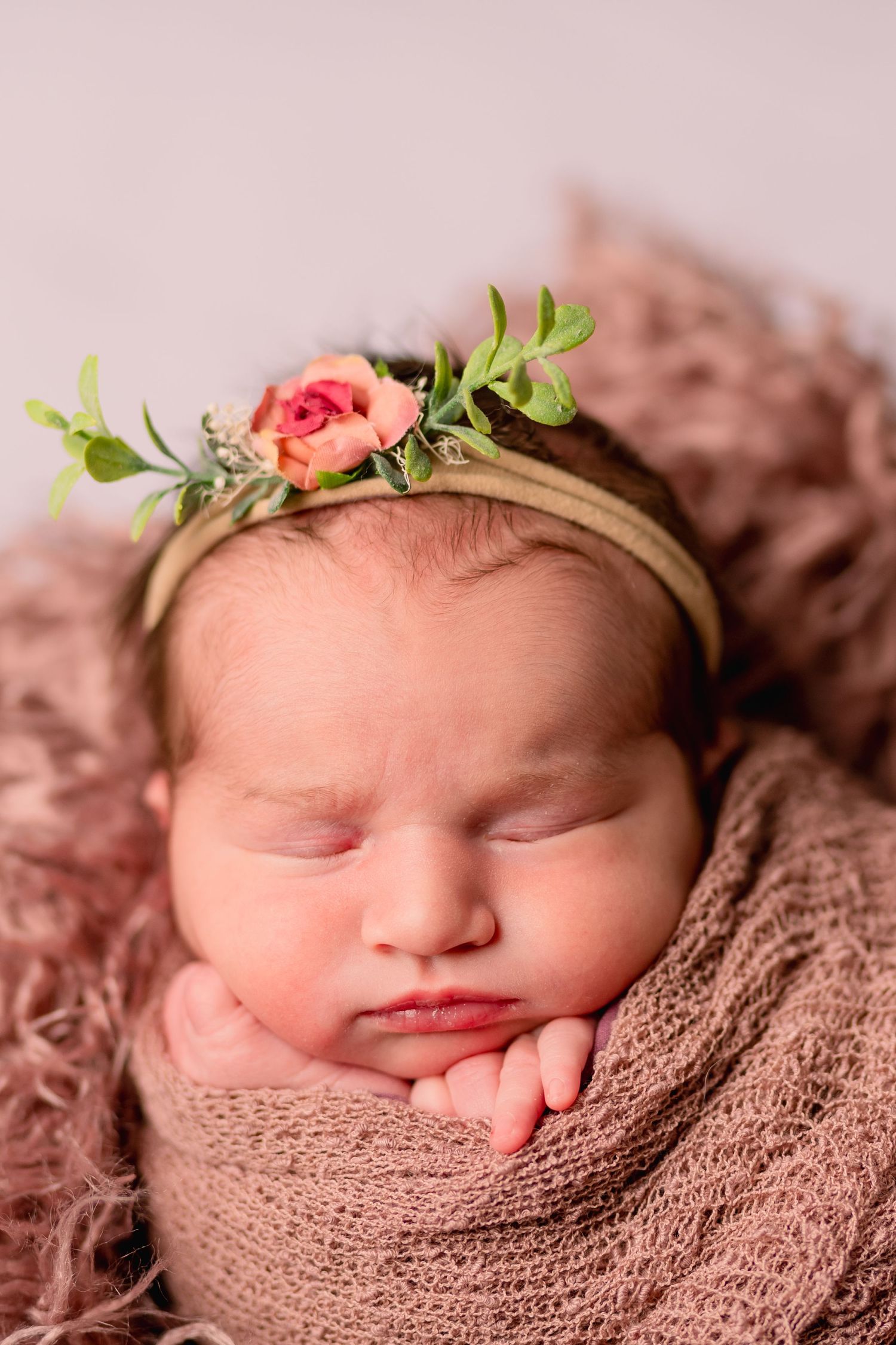 Sleeping newborn wearing floral headband wrapped in mauve blanket for professional portrait.