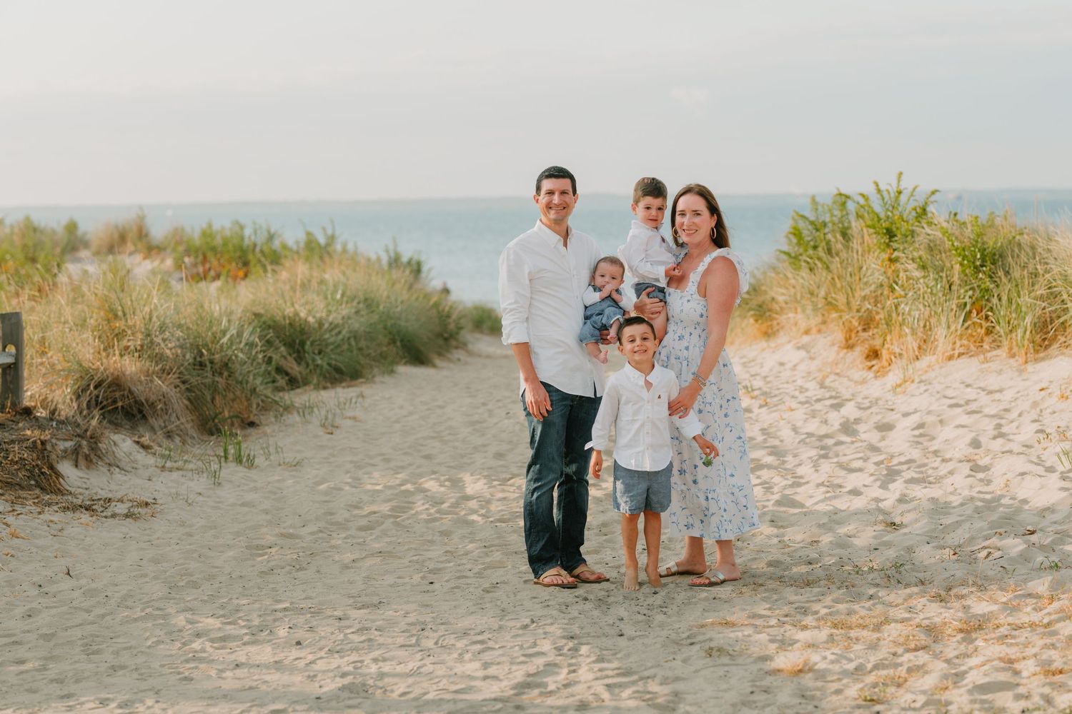 Family portrait on sandy beach path surrounded by coastal grasses at sunset.