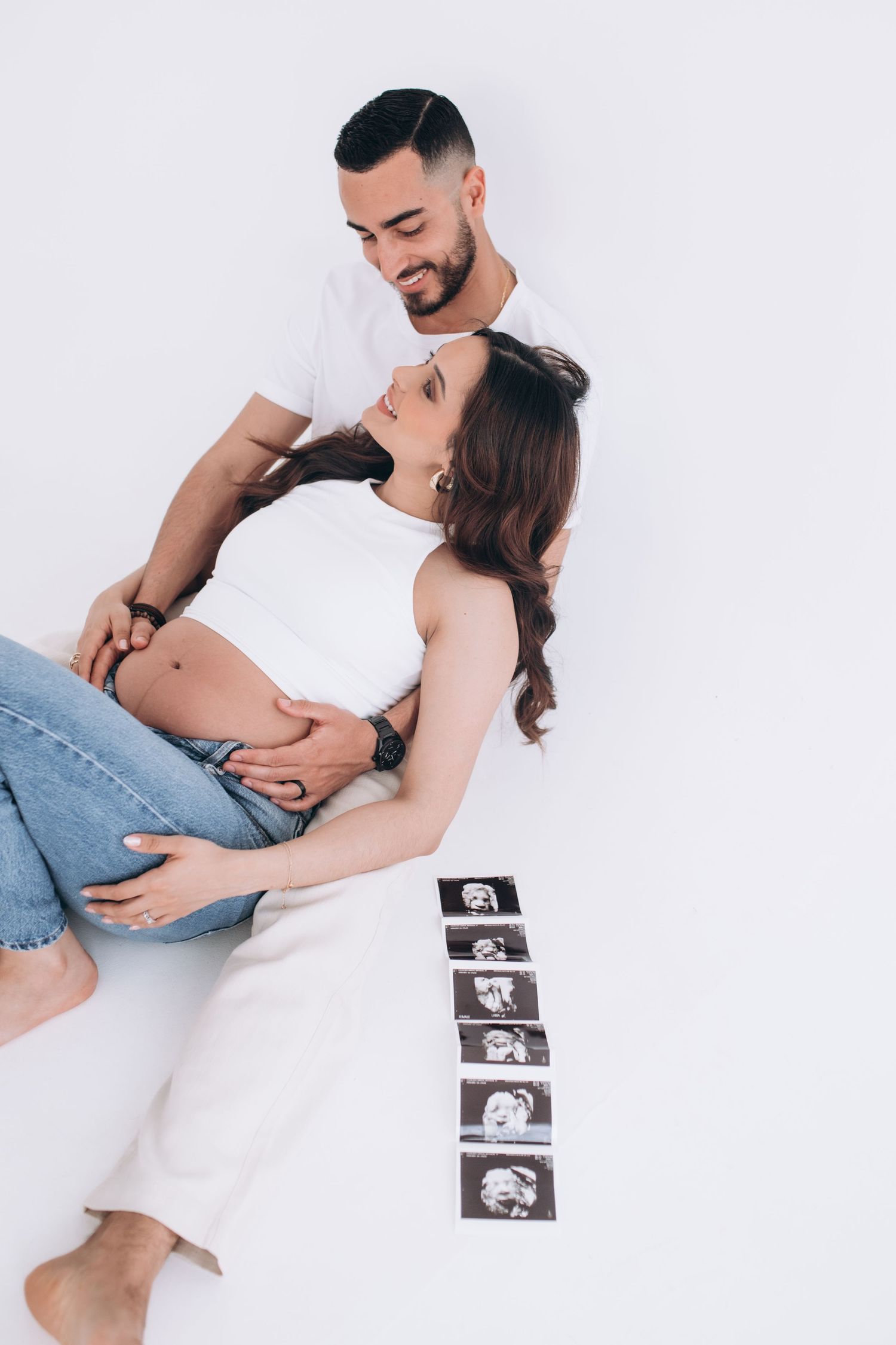 Expectant couple in white outfits lying on floor with ultrasound photos arranged in a row beside them.