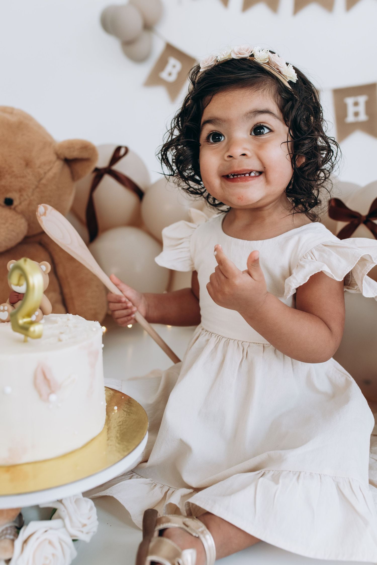 Child in white dress smiling with birthday cake and party decorations in brown and beige tones.