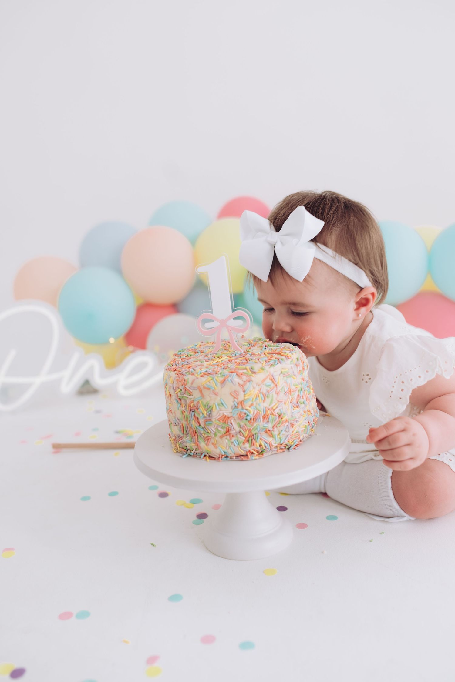 Baby in white dress with bow headband leans toward colorful sprinkle-covered birthday cake with pastel balloons in background.
