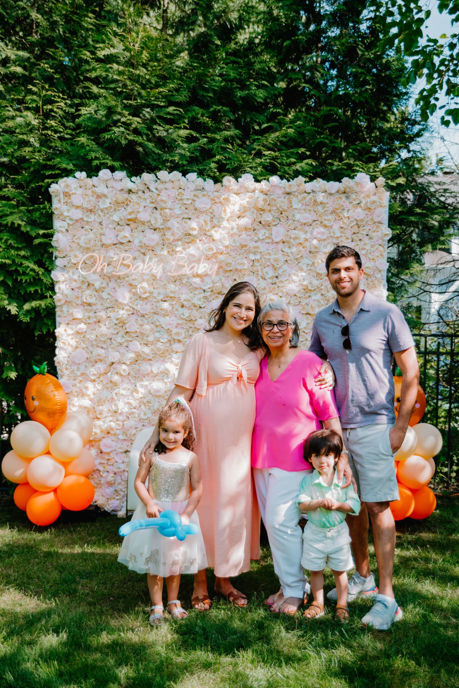 Festive family gathering with floral wall backdrop and orange balloon decorations in an outdoor garden setting.