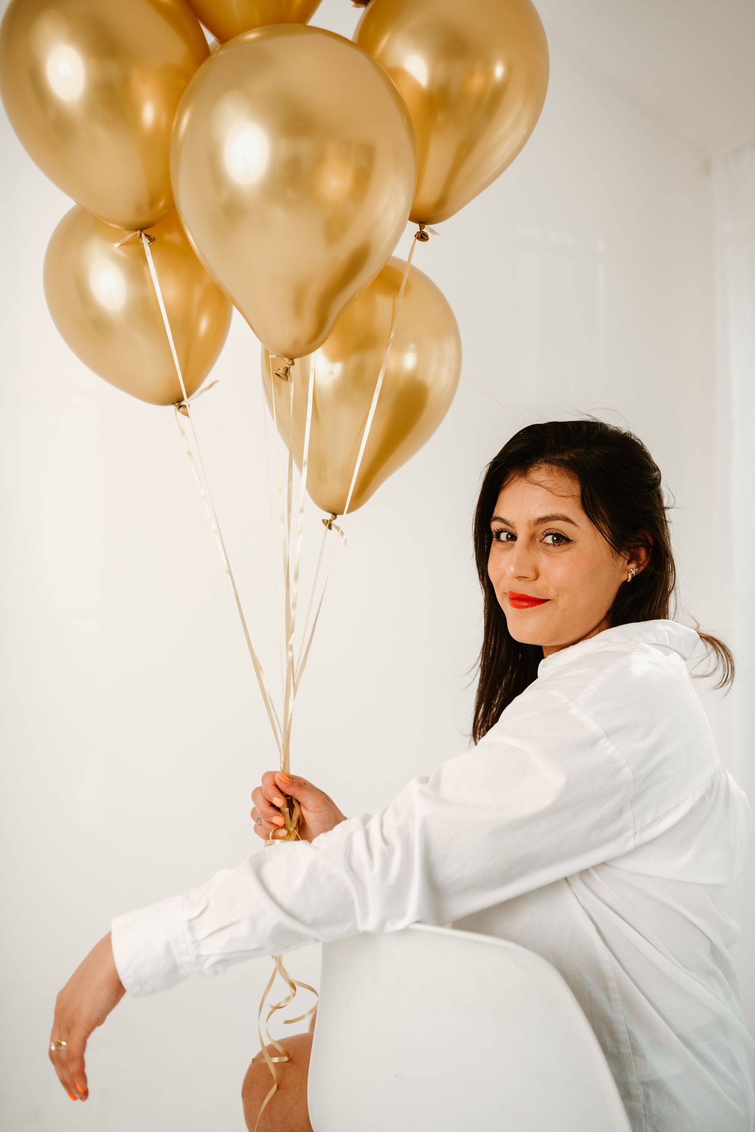 Woman in white shirt poses with gold balloons against white wall.