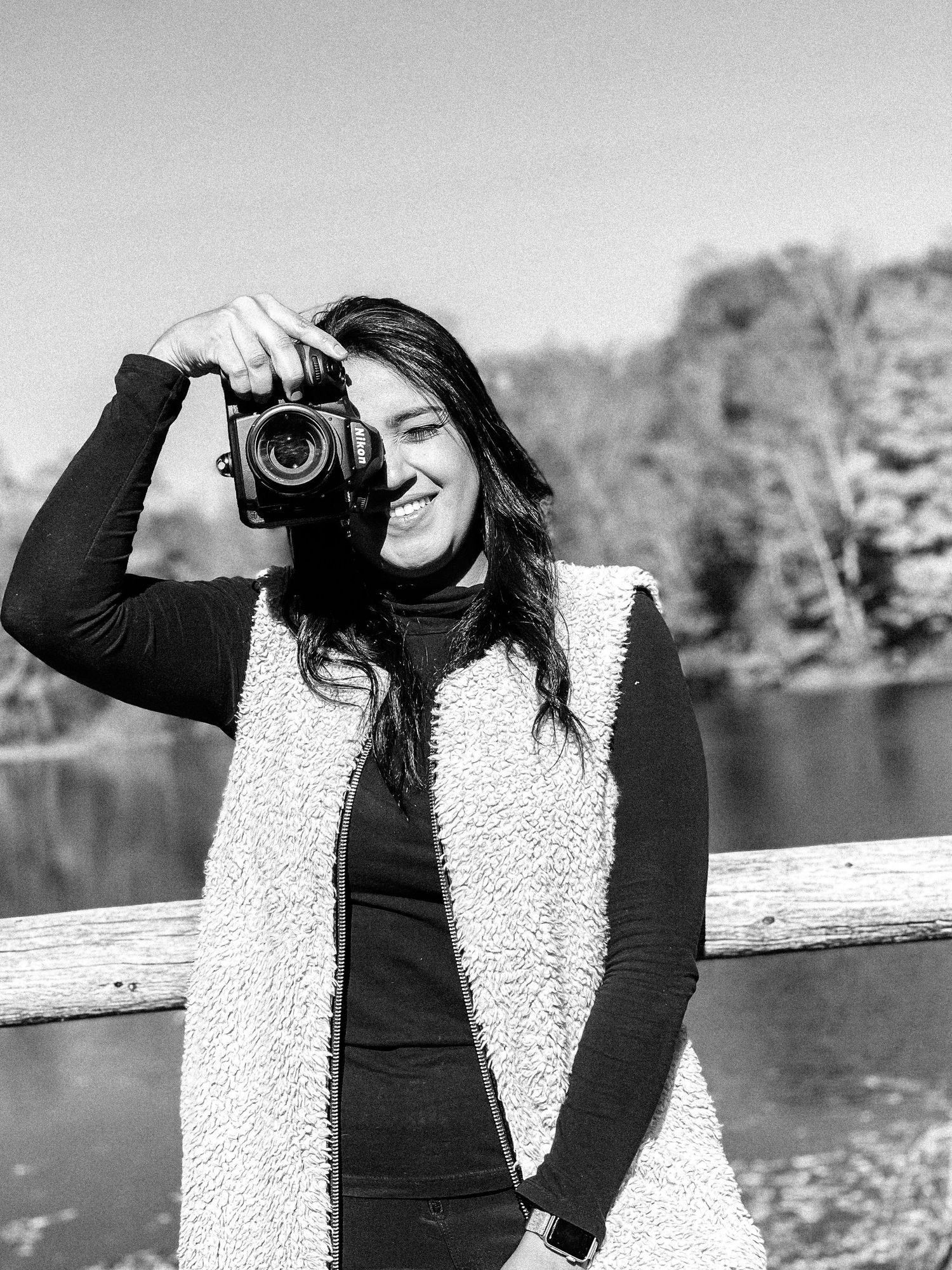 A black and white portrait of a photographer with a camera against a scenic lake backdrop.