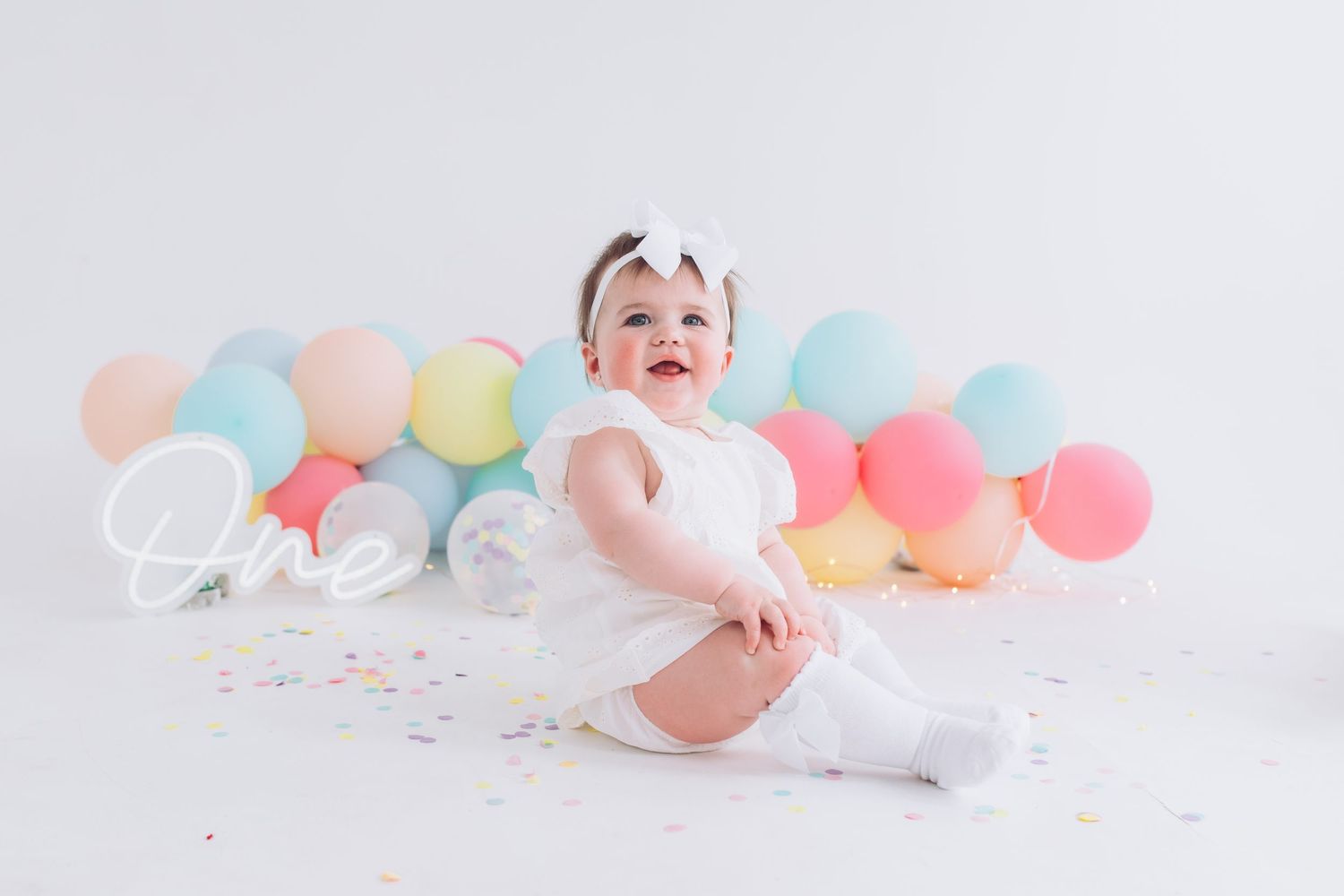Laughing baby in white outfit sits on floor surrounded by pastel balloons and confetti for birthday celebration.