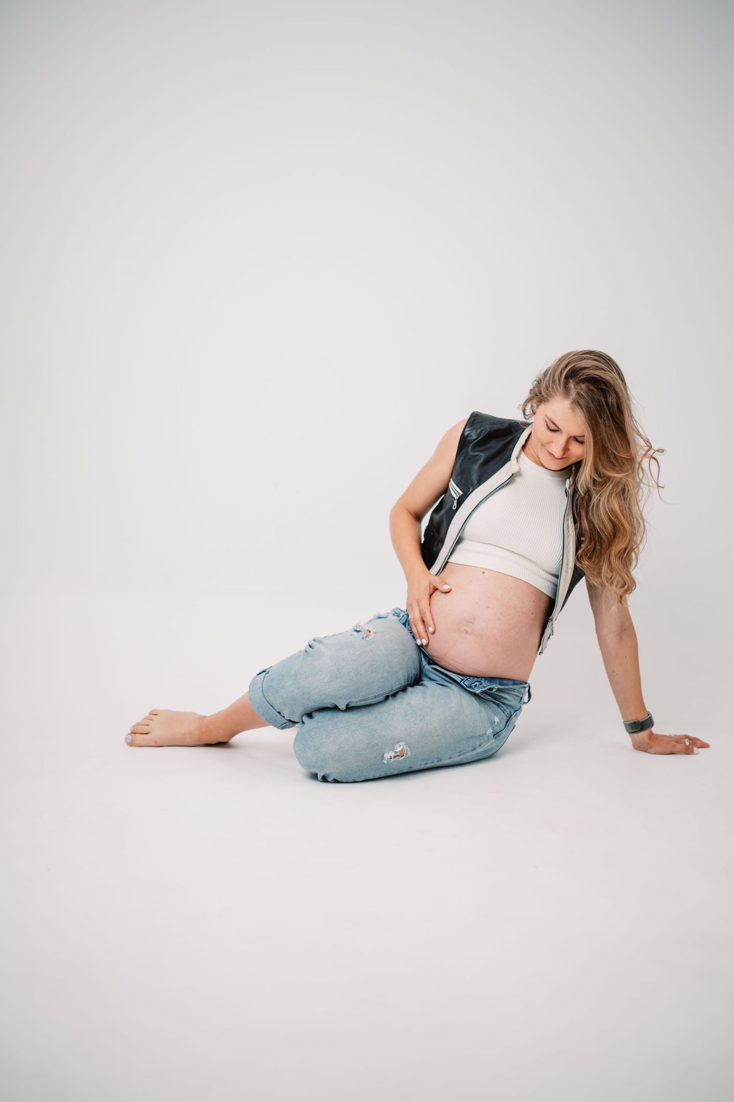 Casual maternity portrait of woman in jeans and vest against white background.
