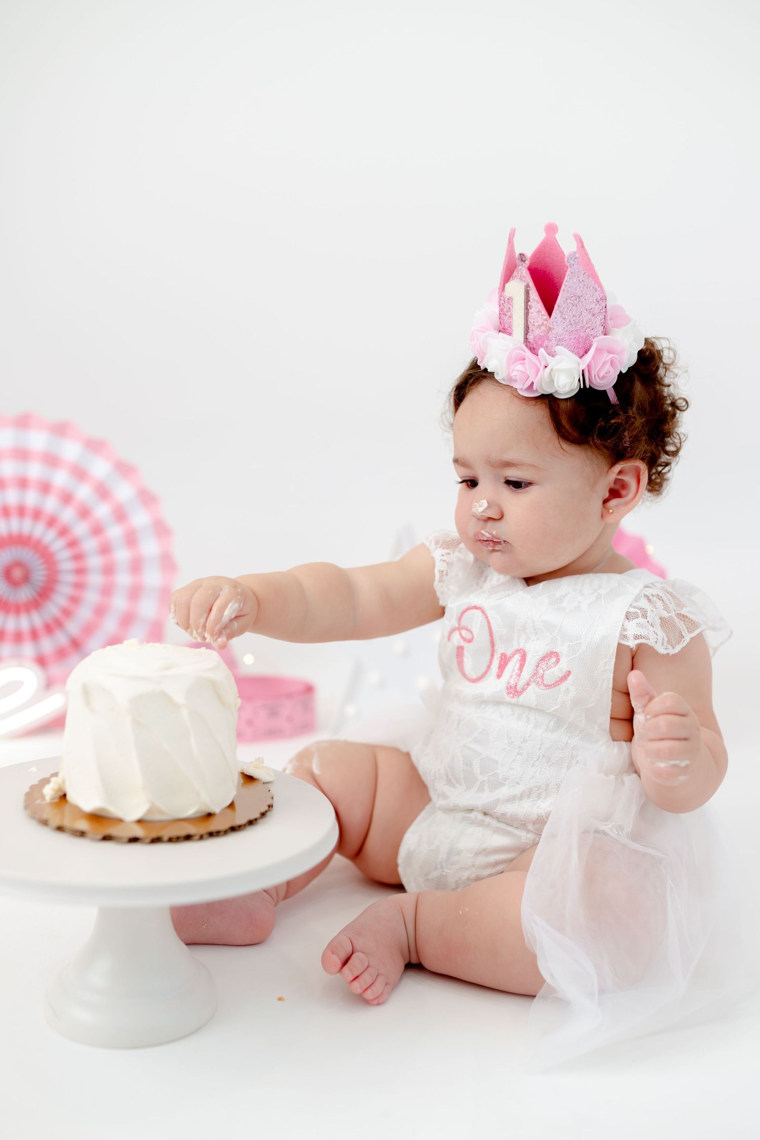 A baby in a white romper and pink crown reaches for a white cake during a first birthday photoshoot.