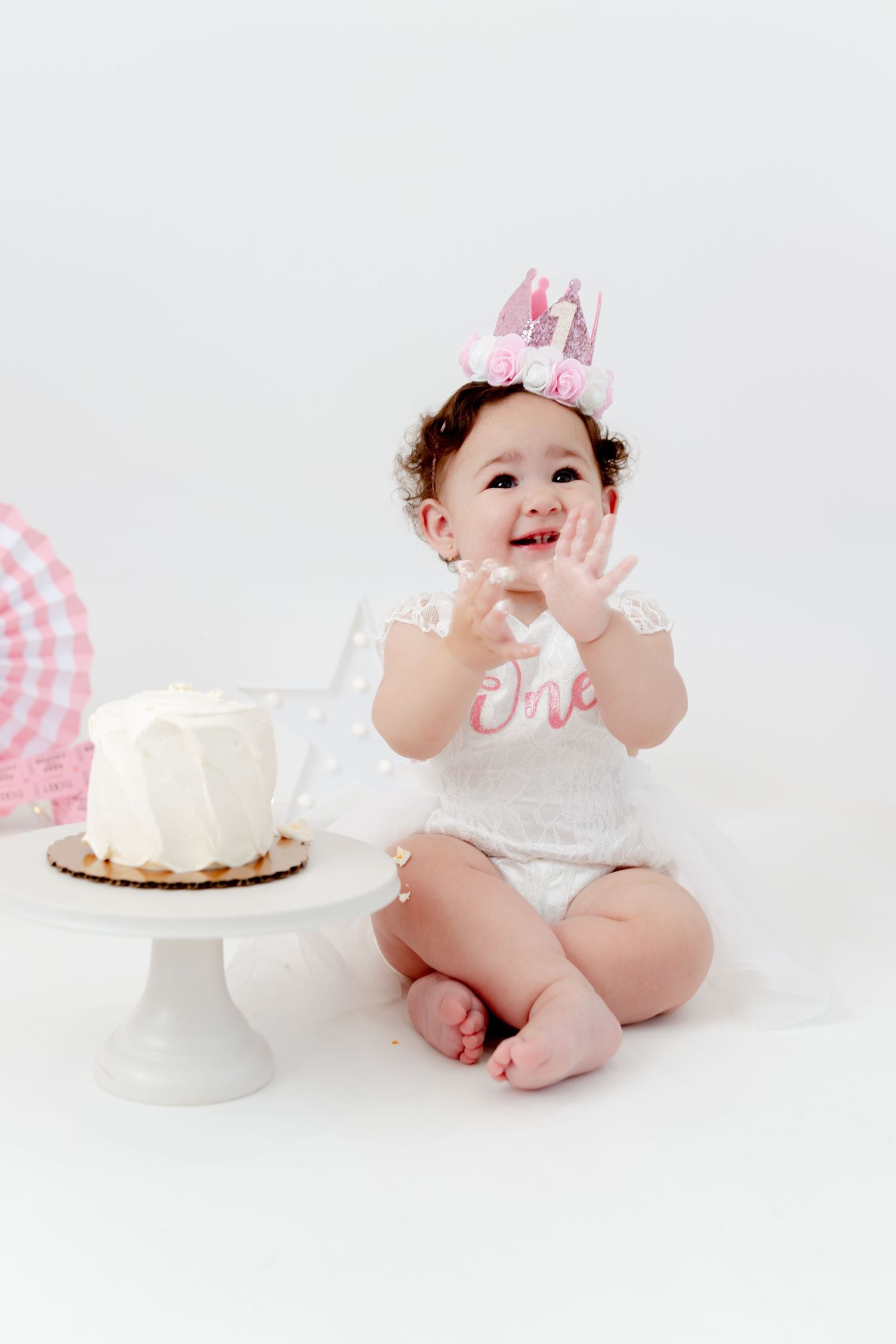 A baby wearing a pink crown and white dress sits next to a white birthday cake.
