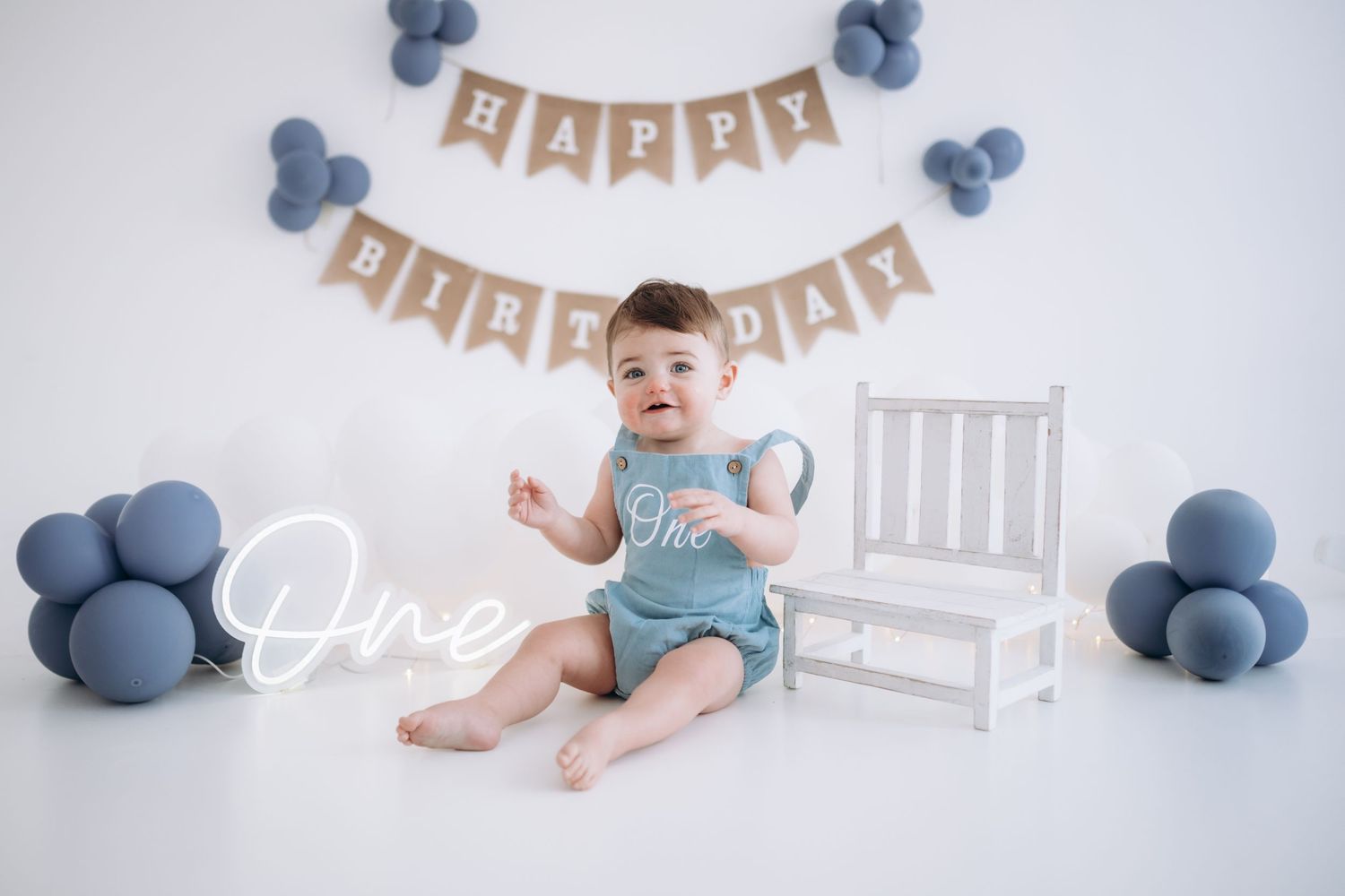 Cake Smash session for baby boy, one year old, Baby in blue outfit sitting by birthday decorations with blue balloons and banner.