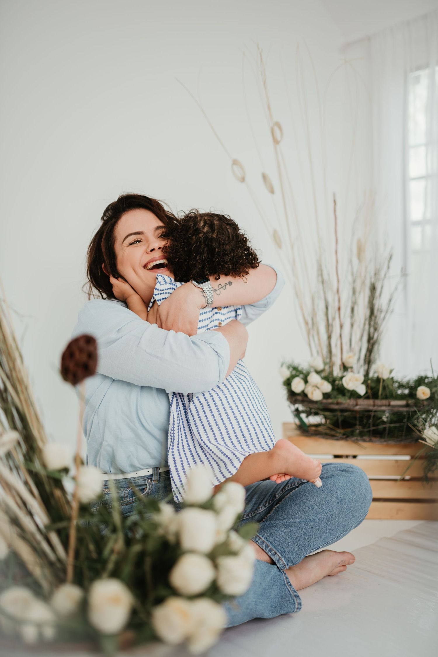 Mother and child share a joyful embrace surrounded by dried flowers and white roses in bright indoor setting.