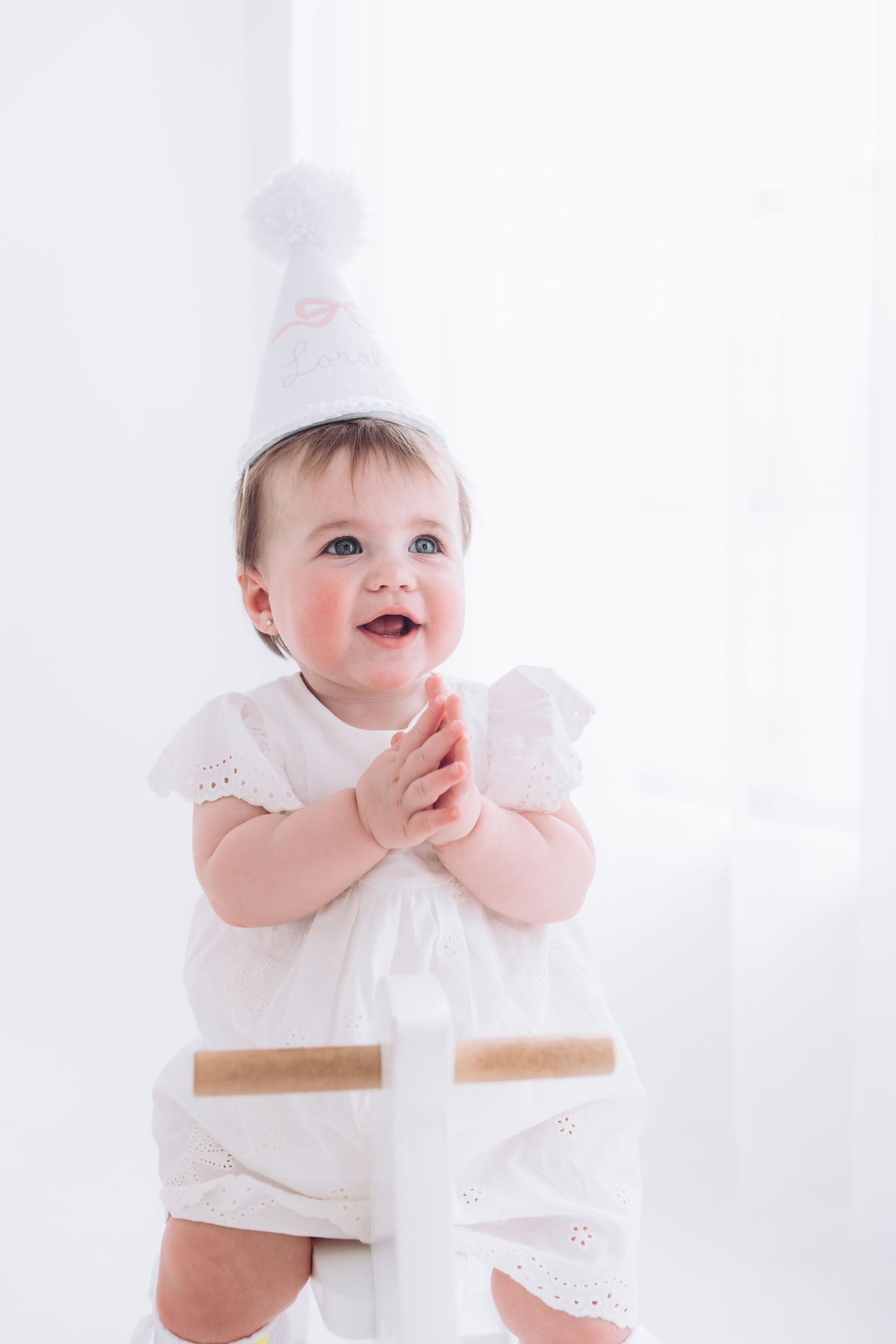 Happy baby in white eyelet lace dress sits against light background with cheerful expression.