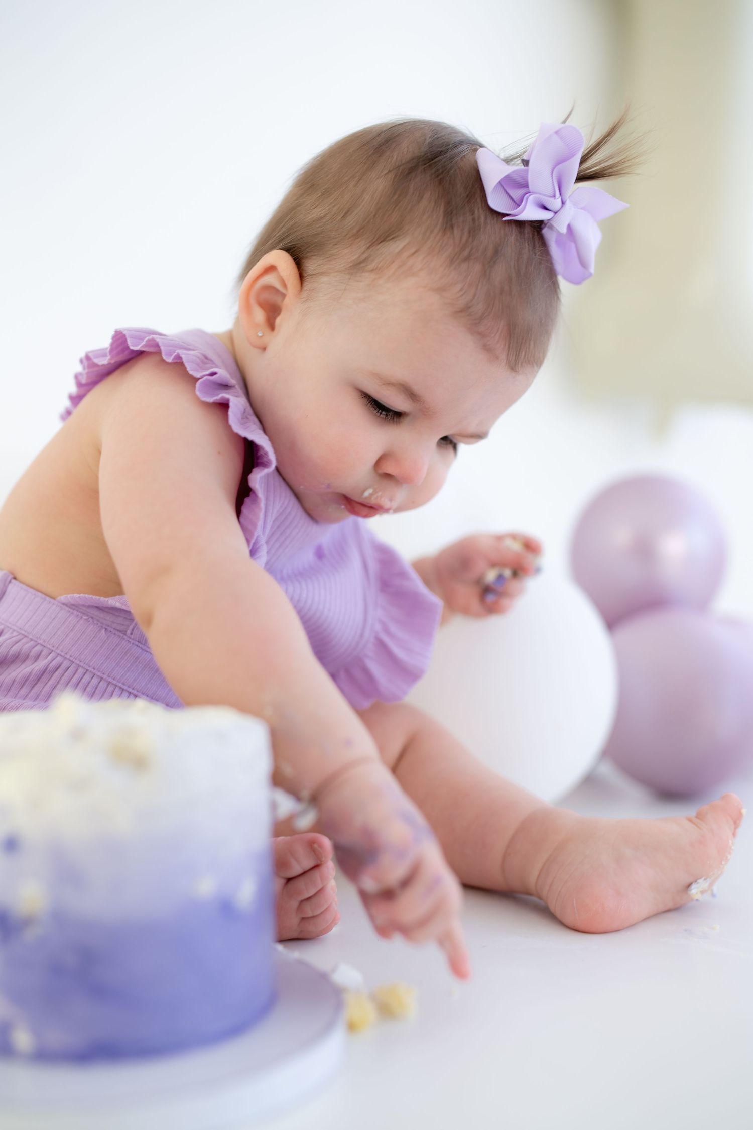 cake smash photography, , Baby in purple outfit with matching bow exploring birthday cake on white background.
