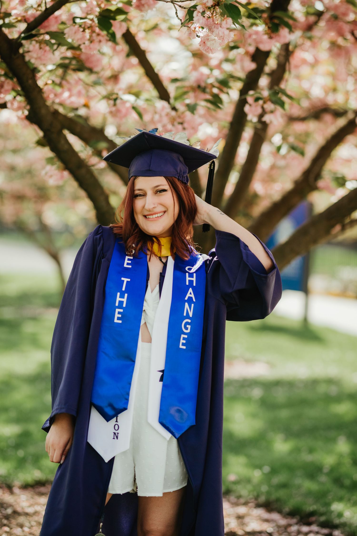 Graduate in navy cap and gown with blue stole smiles under blooming cherry tree.
