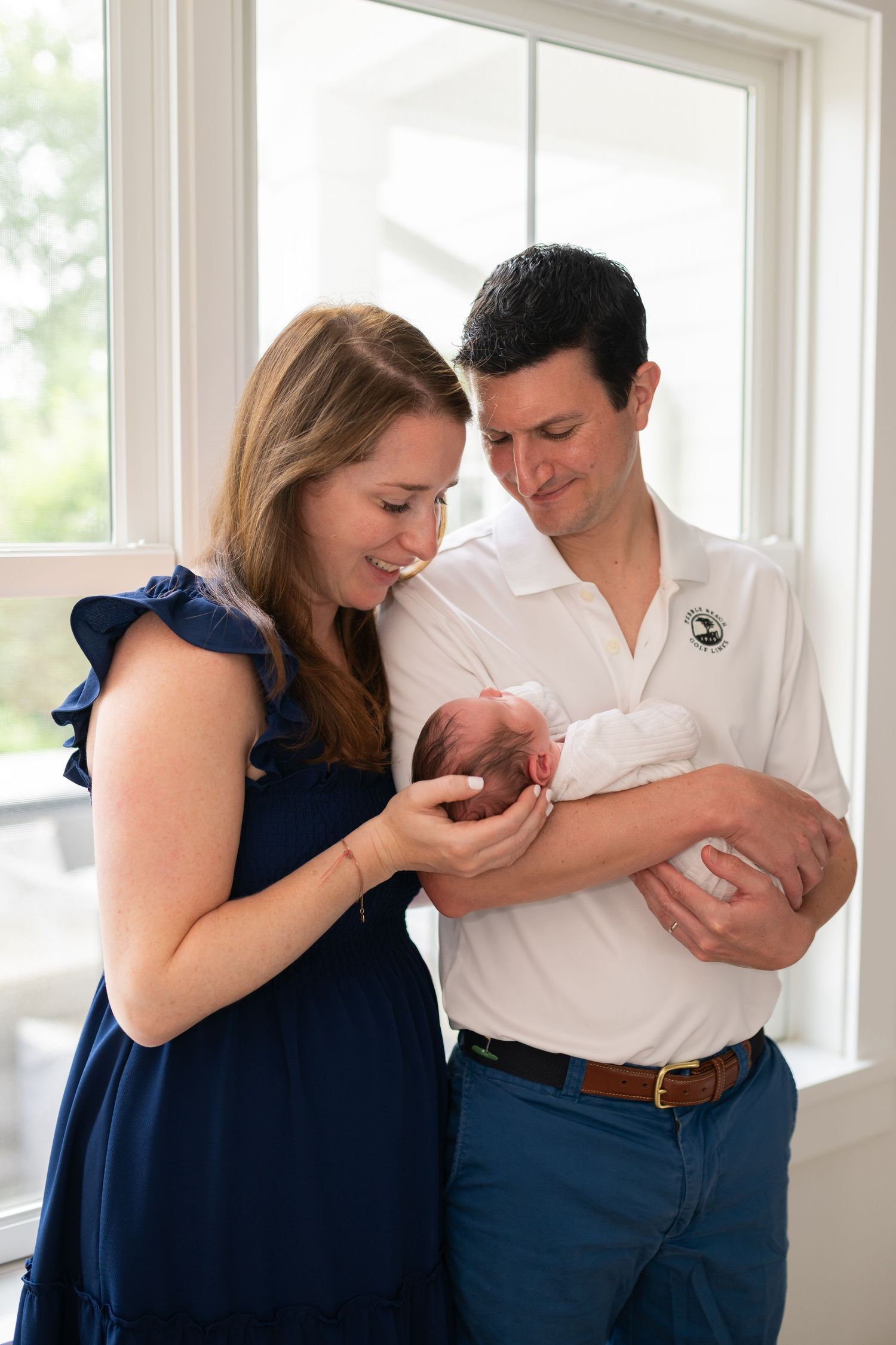 Parents lovingly gaze at their newborn baby near a bright window.