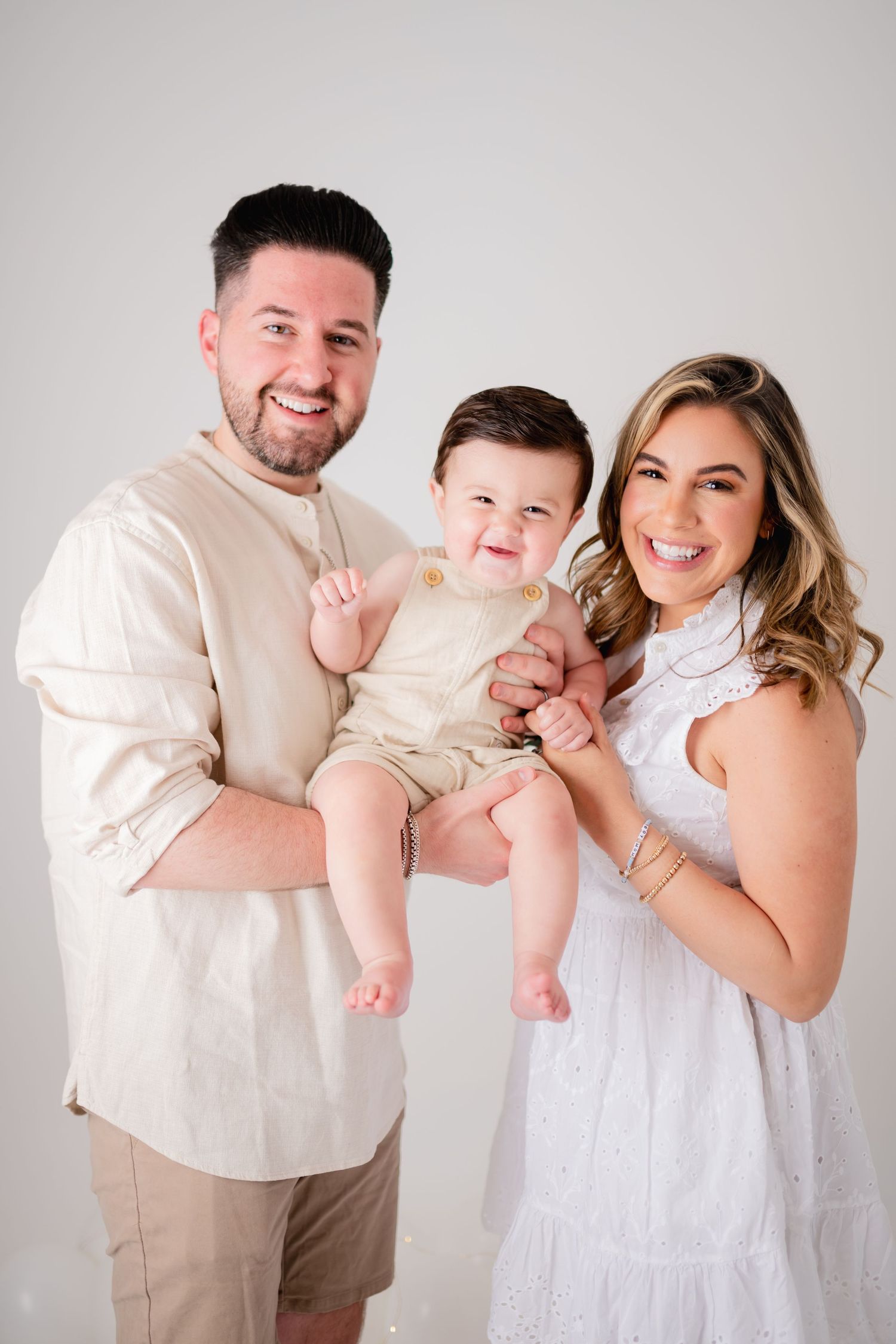 Family of three in coordinating cream and white outfits pose against neutral backdrop.