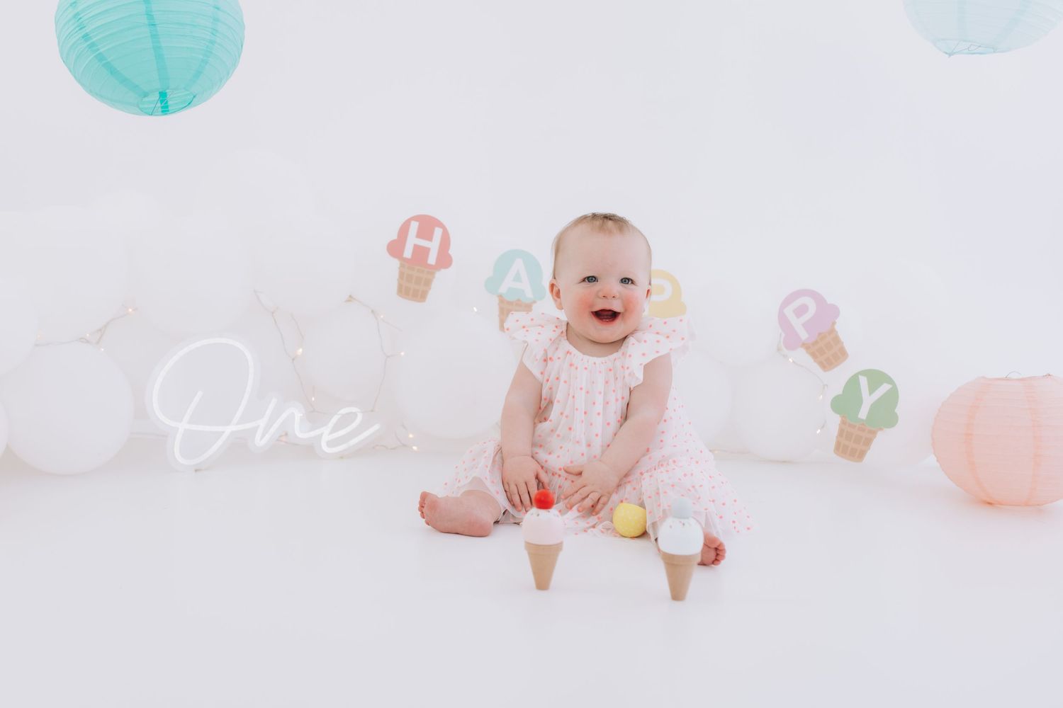 Baby in white lace outfit sits surrounded by pastel balloons and decorations against a white backdrop.