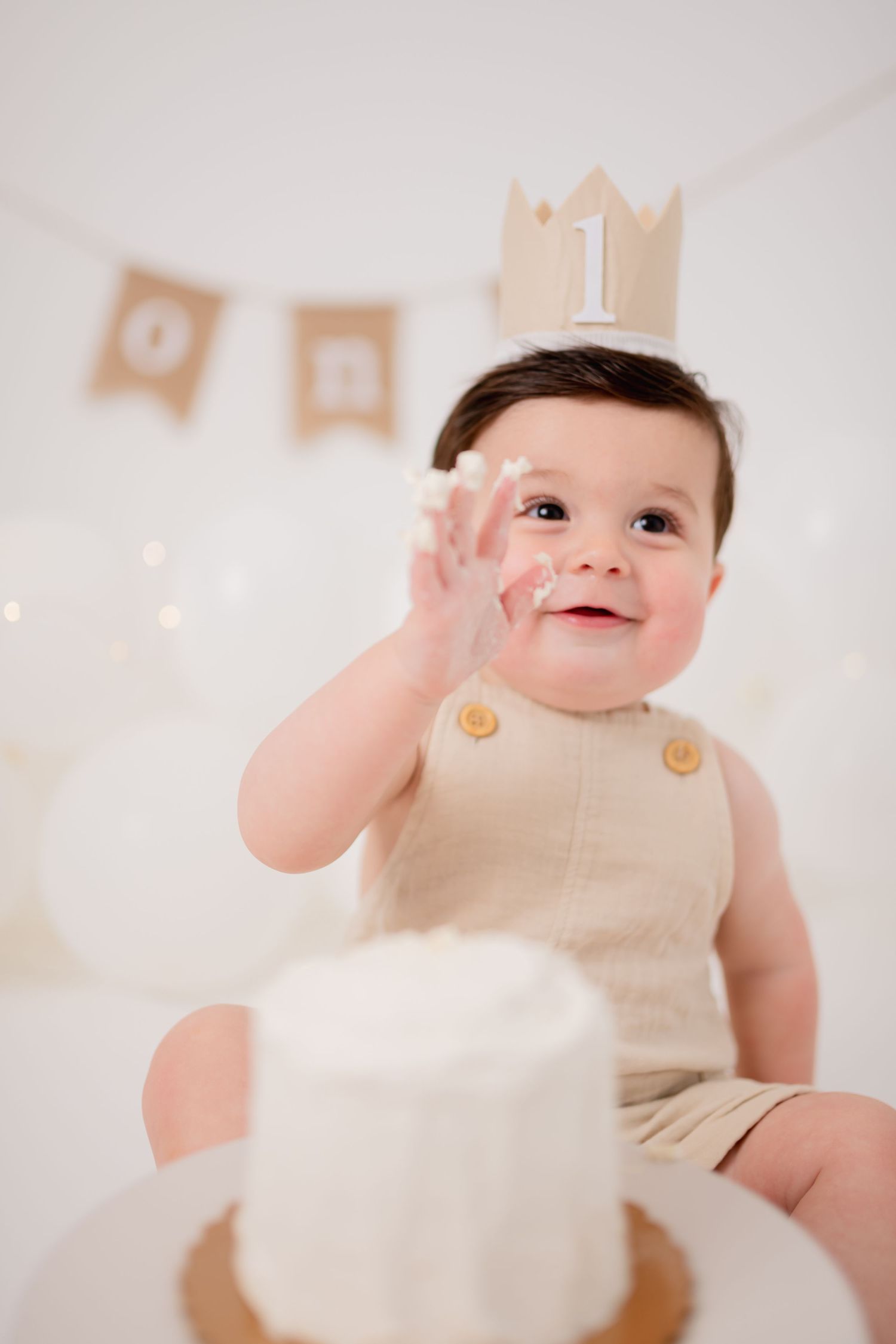 Baby in beige romper sits by birthday cake with crown decoration and banner.