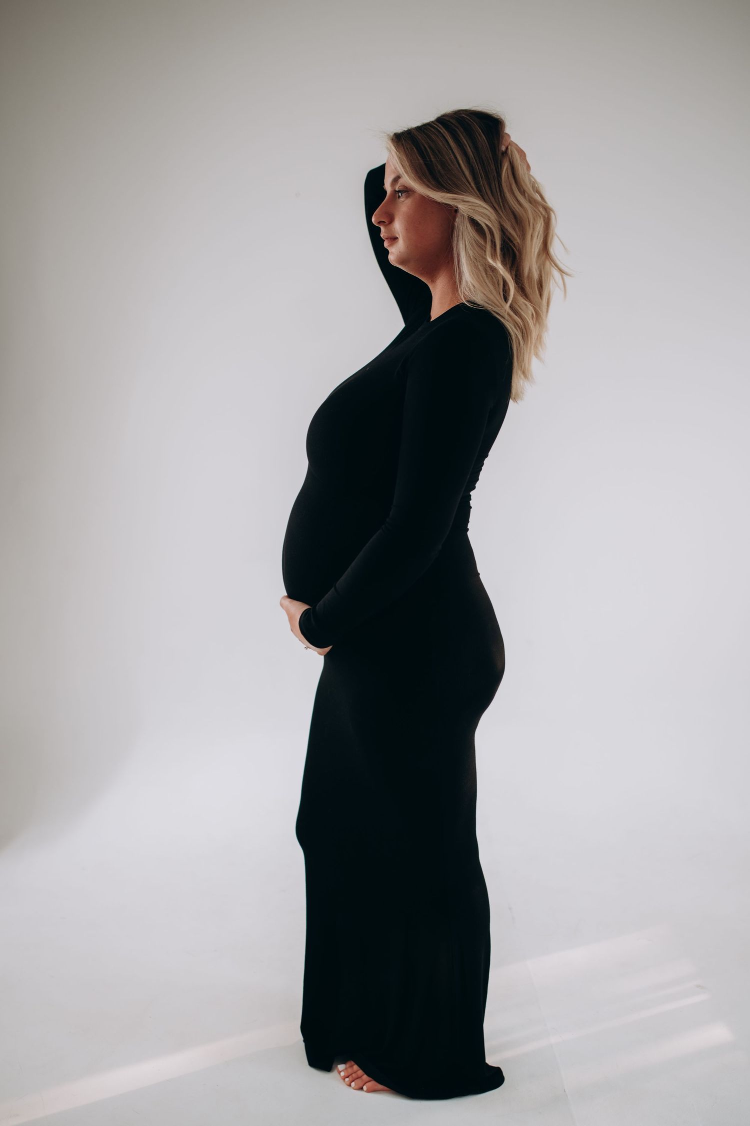 Side profile of an expectant mother wearing a long black dress in a minimalist studio setting against white background.