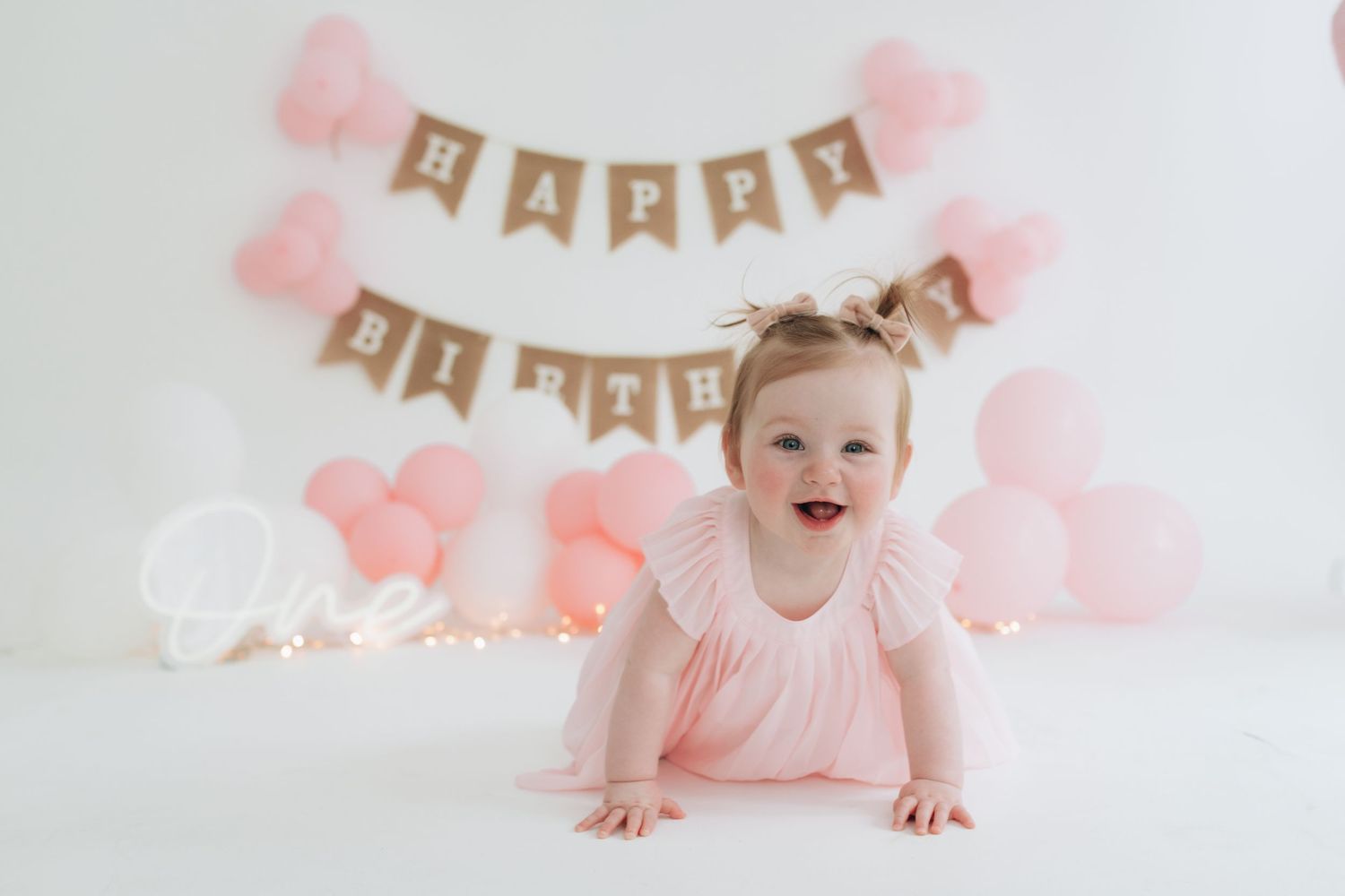 Sweet first birthday setup with pink balloons, bunting banner and small figure in pink dress crawling on white floor.