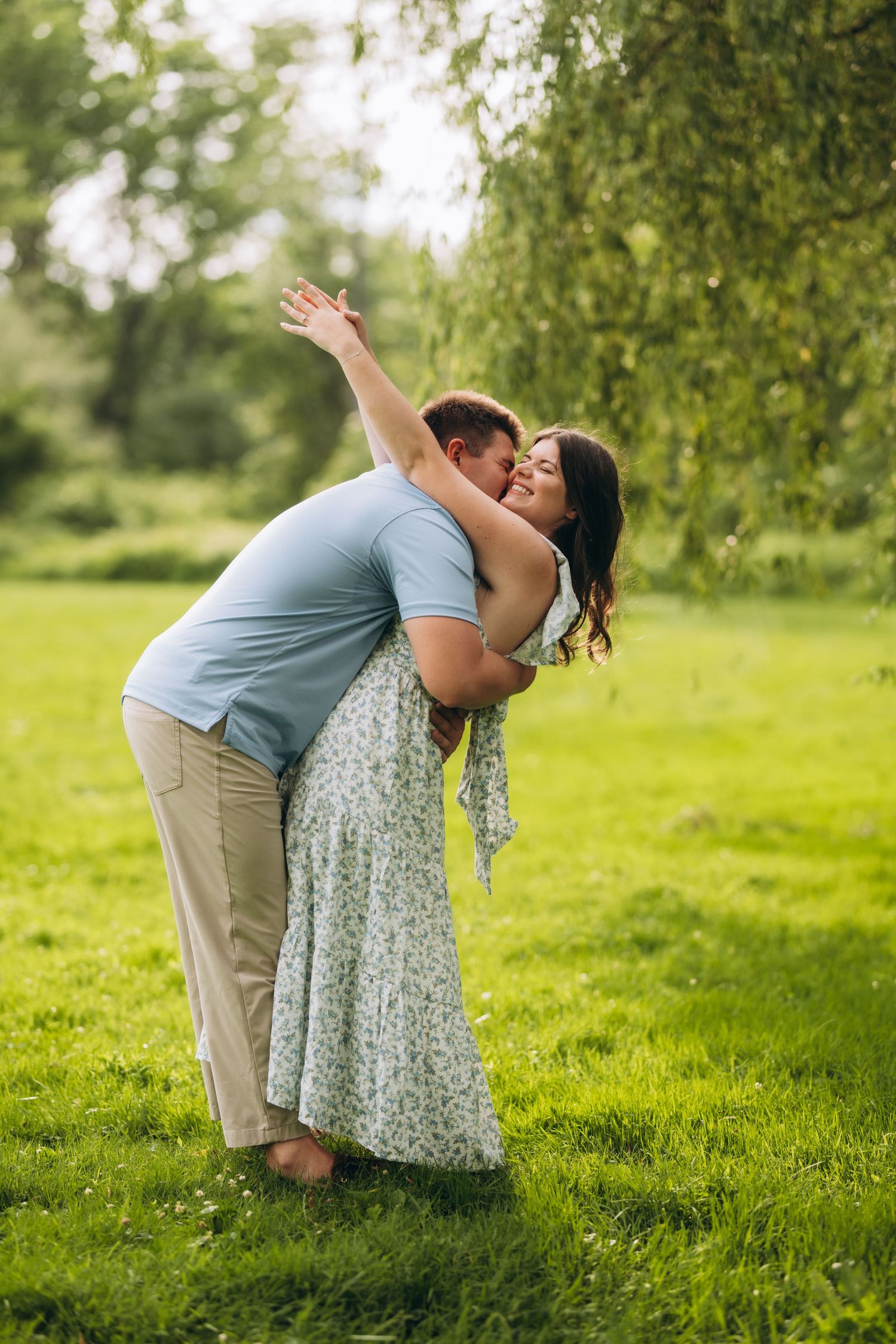 A couple embraces in a romantic dip pose on lush green grass under drooping willow tree branches on a sunny day.