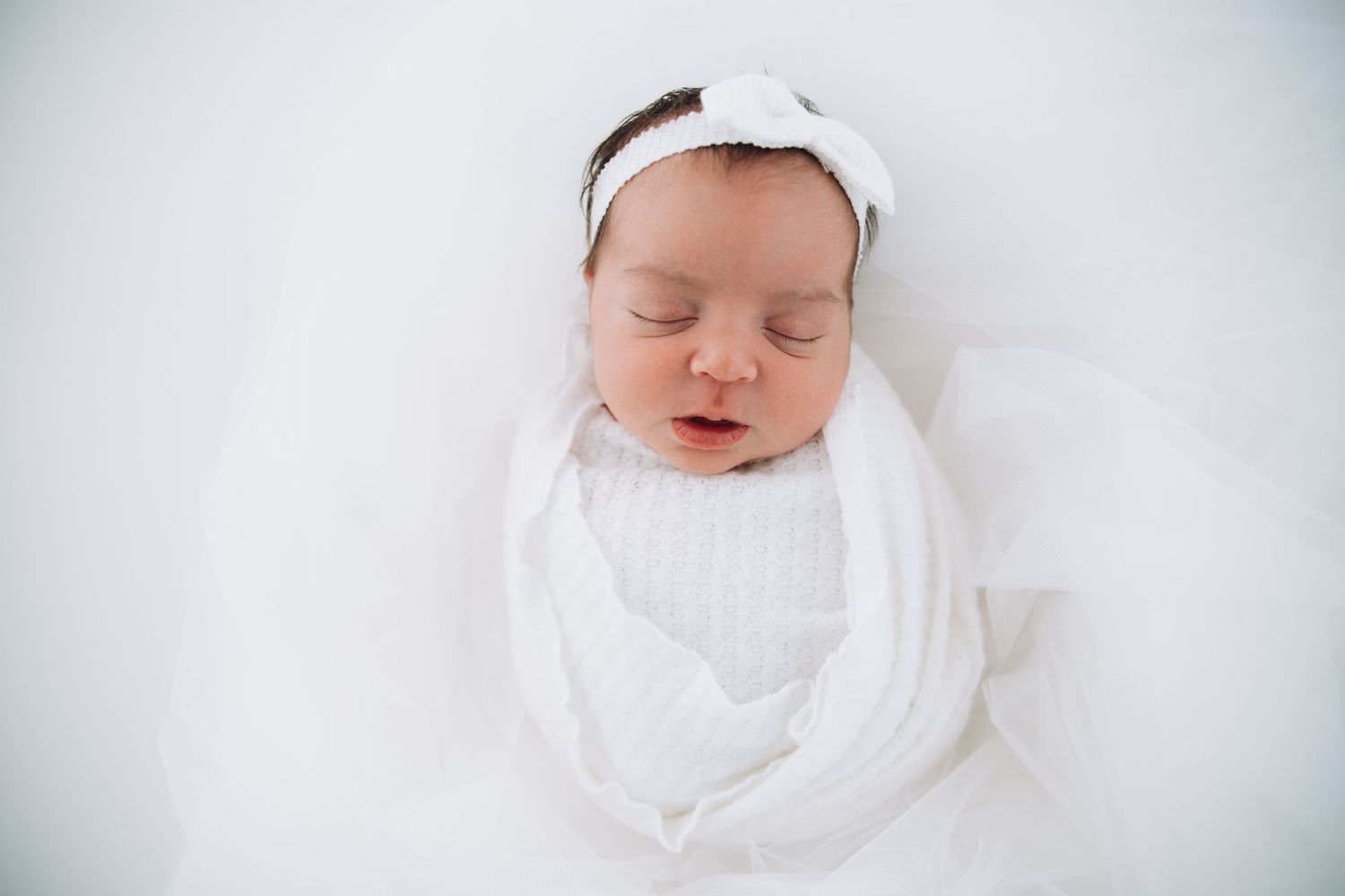 A peaceful sleeping newborn wrapped in white swaddle cloth with a matching white headband on soft fabric background.