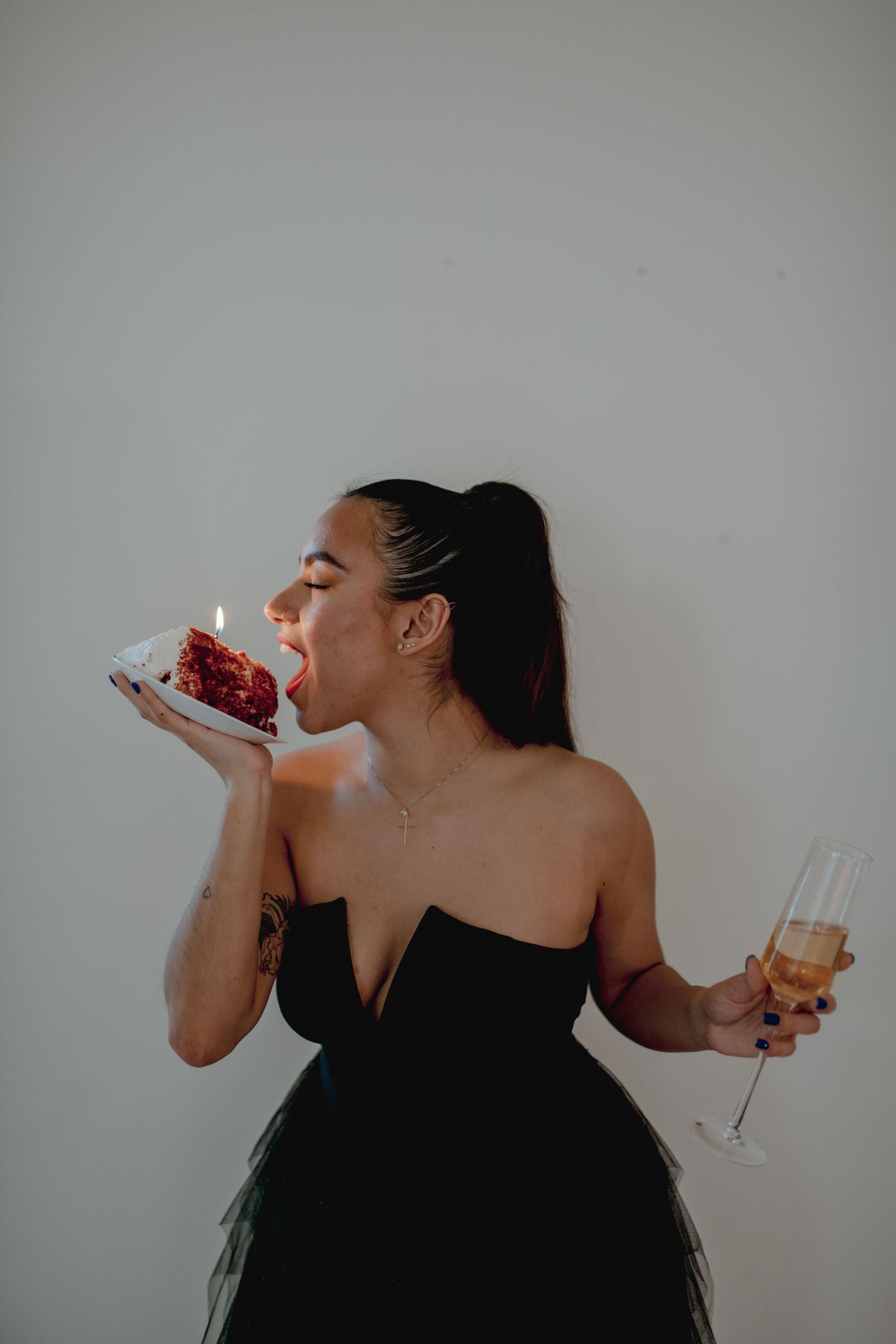 Woman in black dress takes bite of red cake while holding champagne.