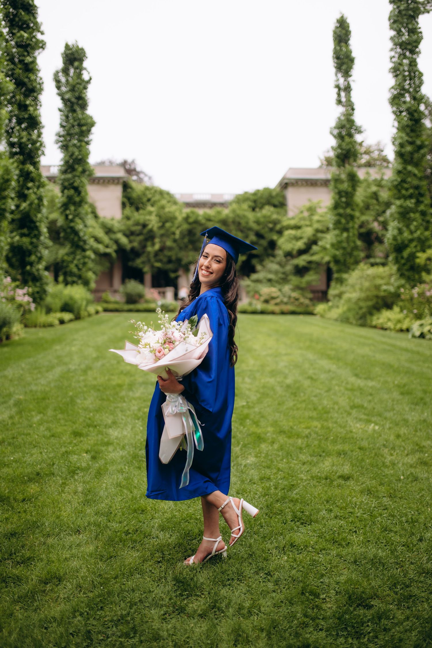 Graduate in royal blue cap and gown leaps joyfully on a grassy lawn during a celebratory photoshoot.