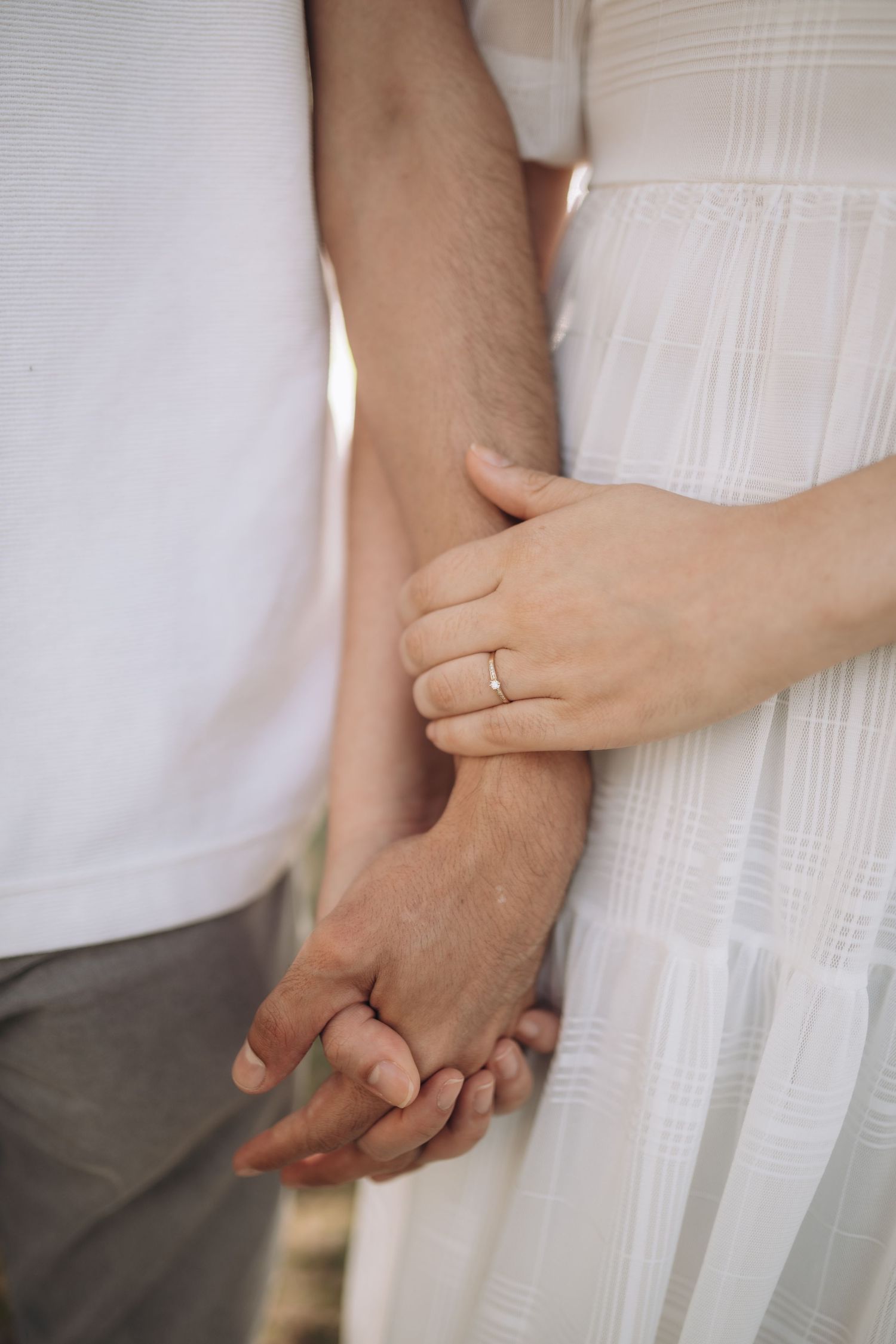 Close up of couple holding hands with engagement ring visible against white dress and casual attire.