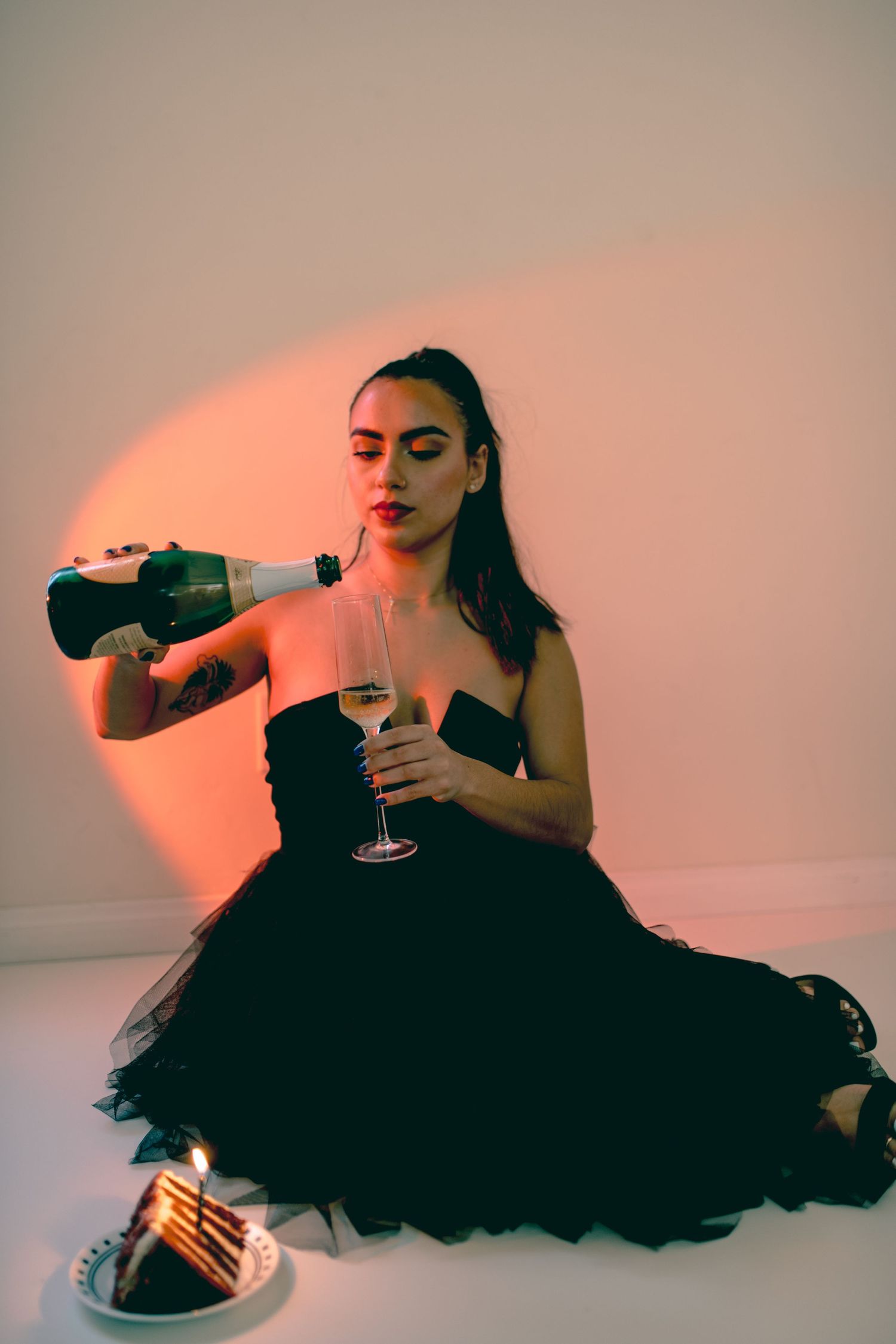 A woman in black tulle dress pours champagne while sitting on the floor.