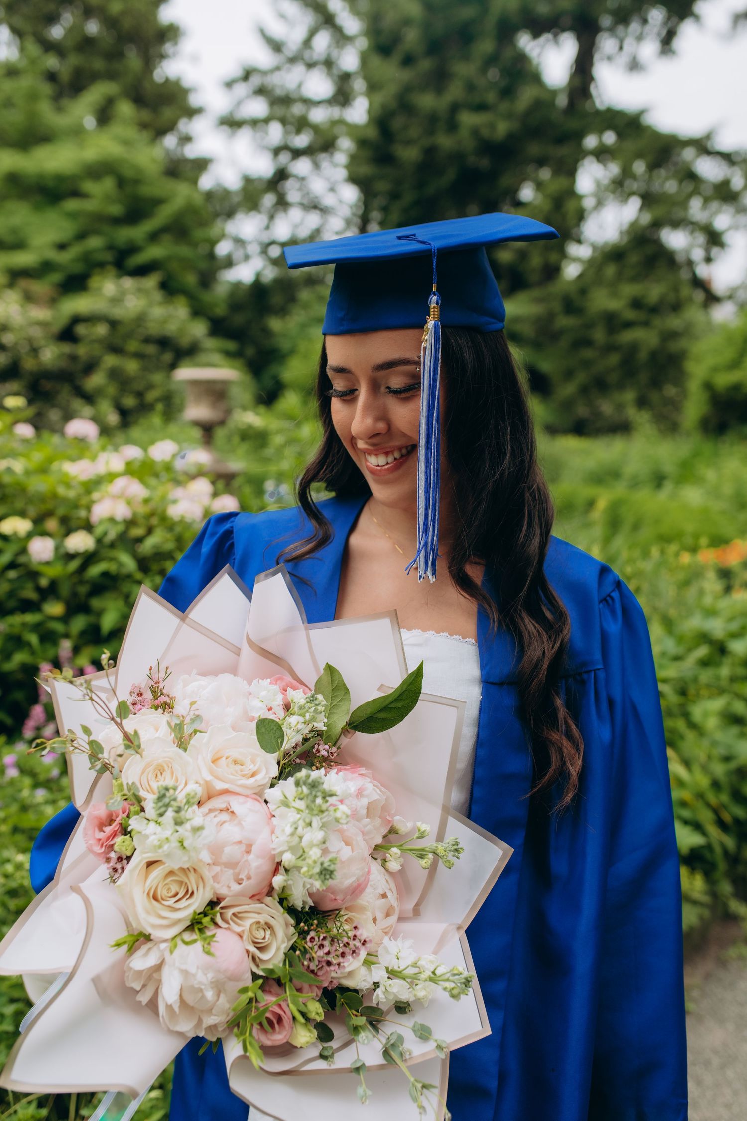 Graduate in royal blue cap and gown holds elegant bouquet of pink and white roses in garden setting.