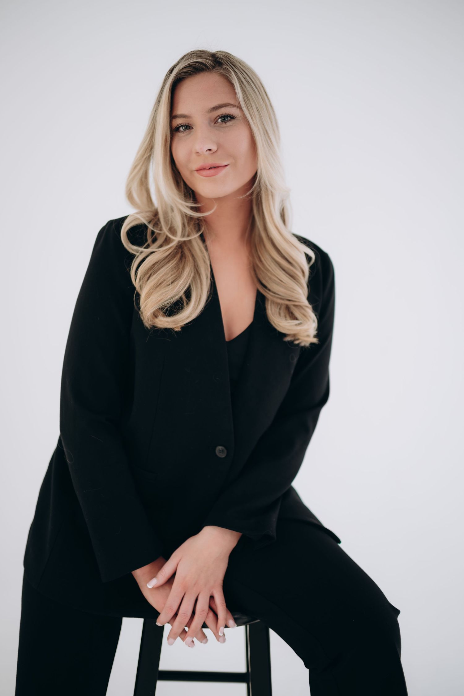 Professional headshot in black business attire seated on stool against white background.