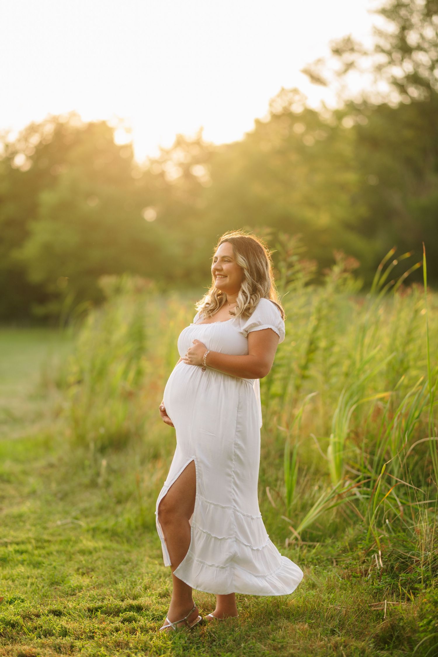 Expectant mother in flowing white dress poses in golden sunlight amid tall grasses.