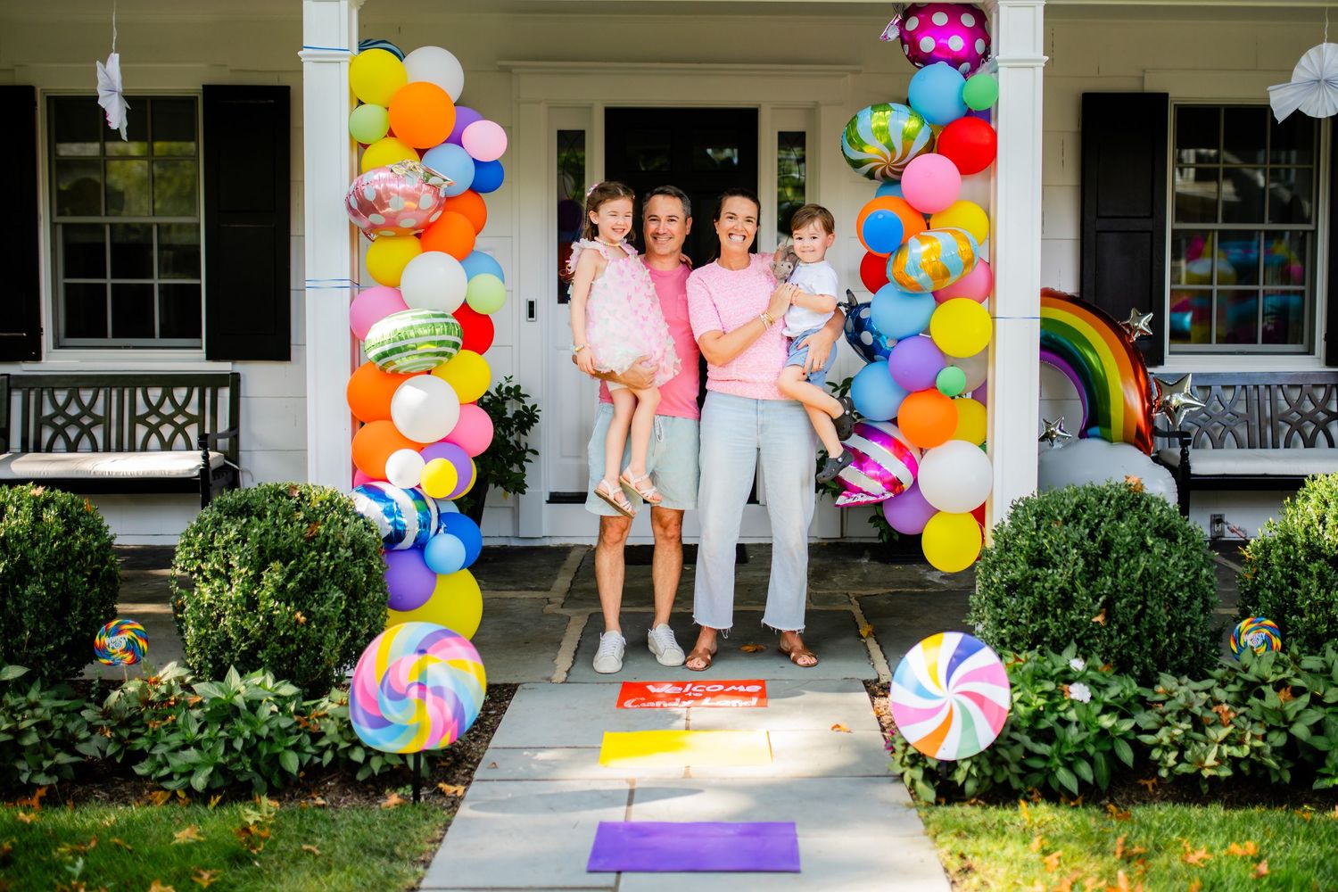 Family on porch with candy-themed decorations and rainbow balloon arch.