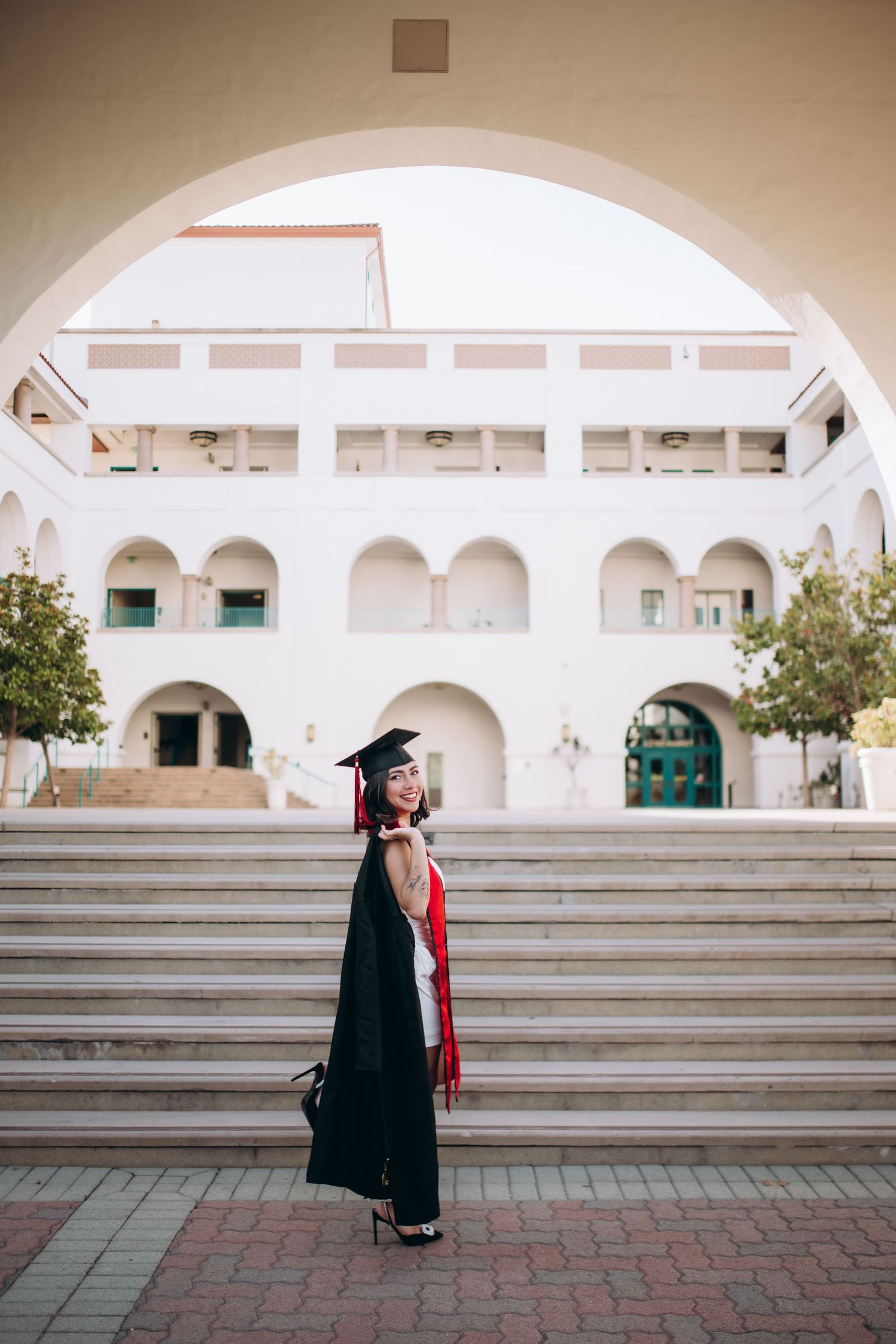Graduate in cap and gown standing on steps of Spanish colonial architecture building.