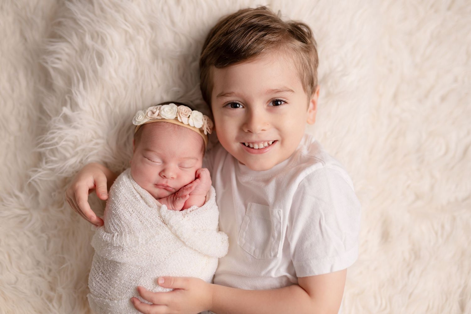 Two siblings posing on white fur rug with newborn wrapped in white.