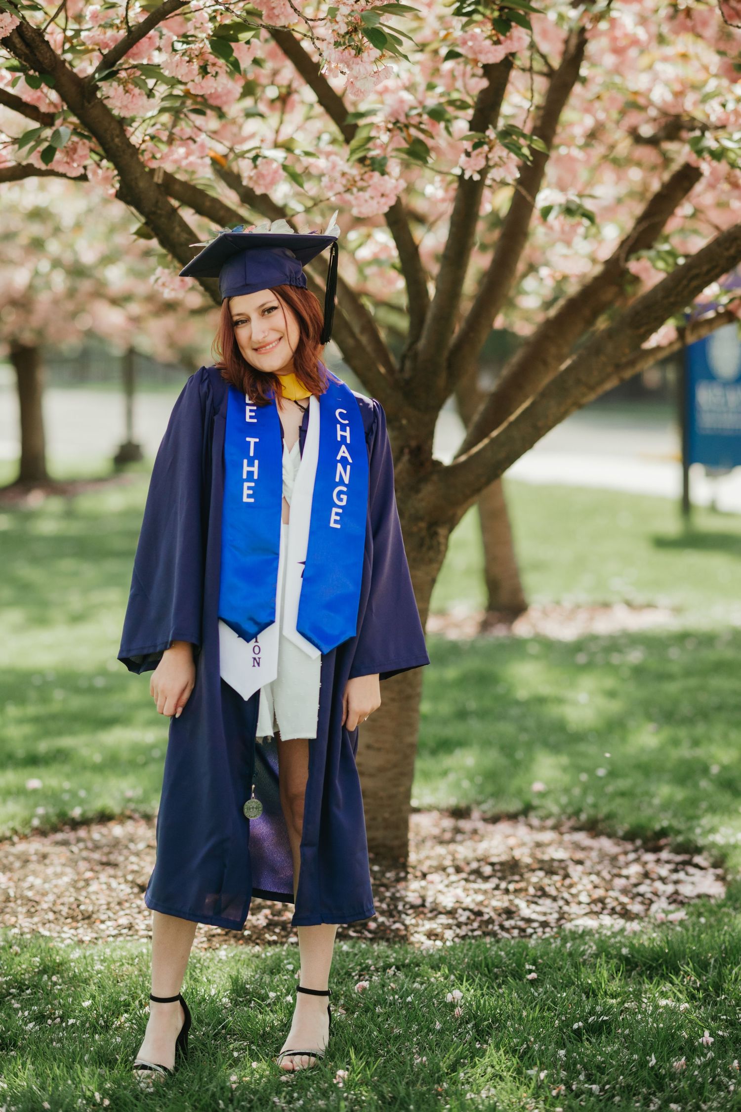 Graduate in navy gown and blue stole poses under blooming cherry tree.