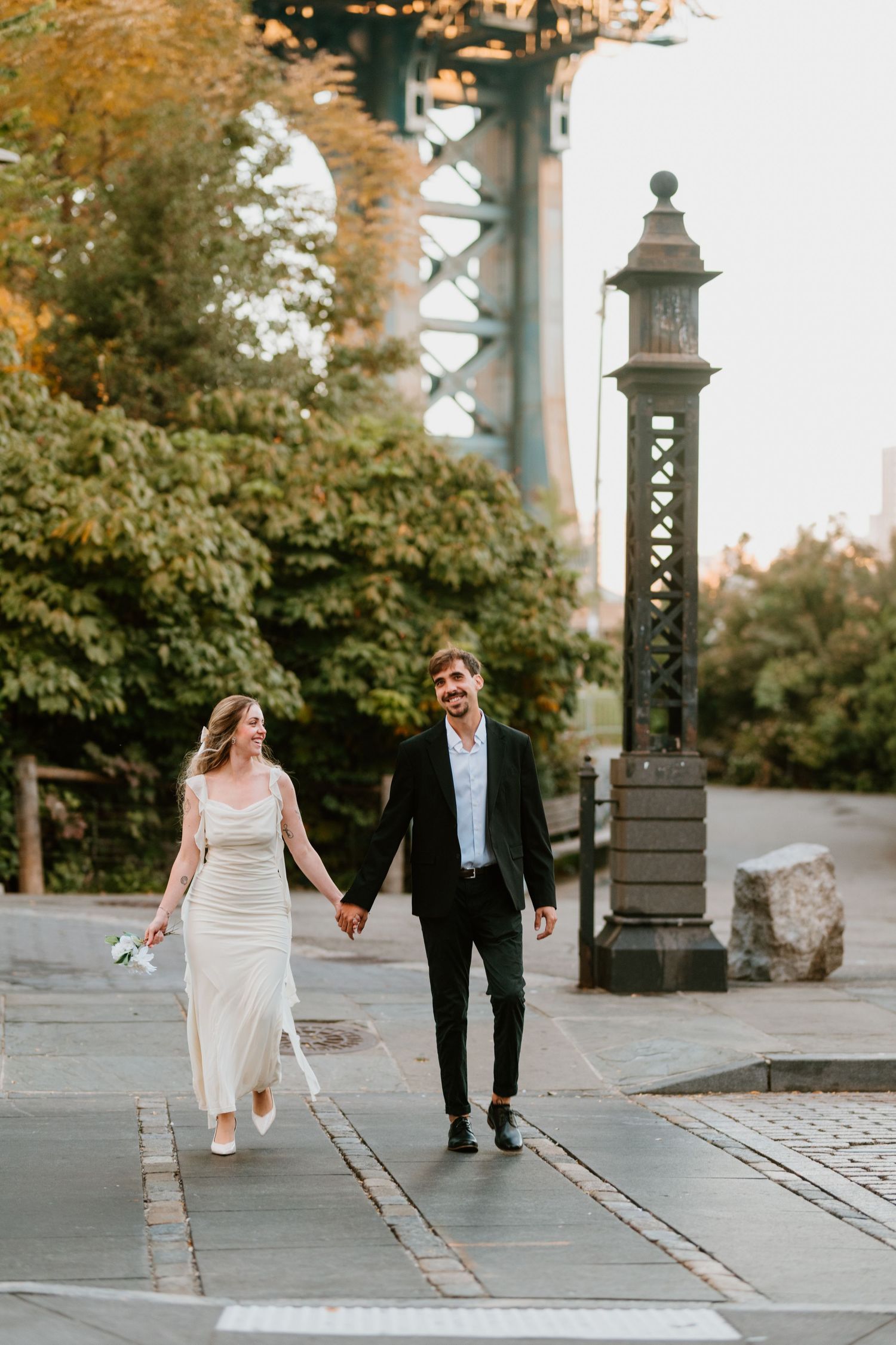 Newlywed couple walking hand in hand near a vintage lamppost and bridge.