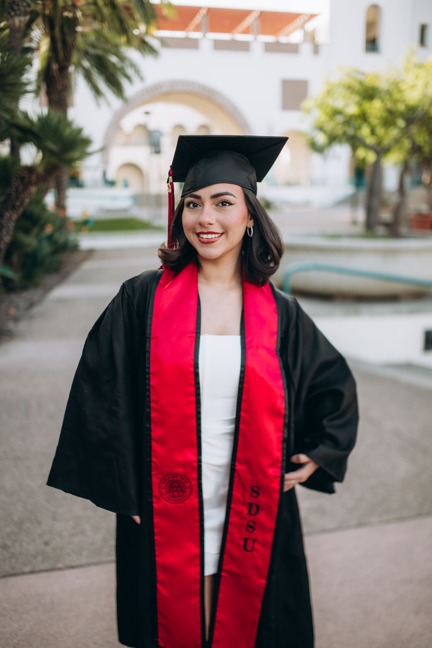 Graduate wearing black cap and gown with red stole stands in front of mission-style building.