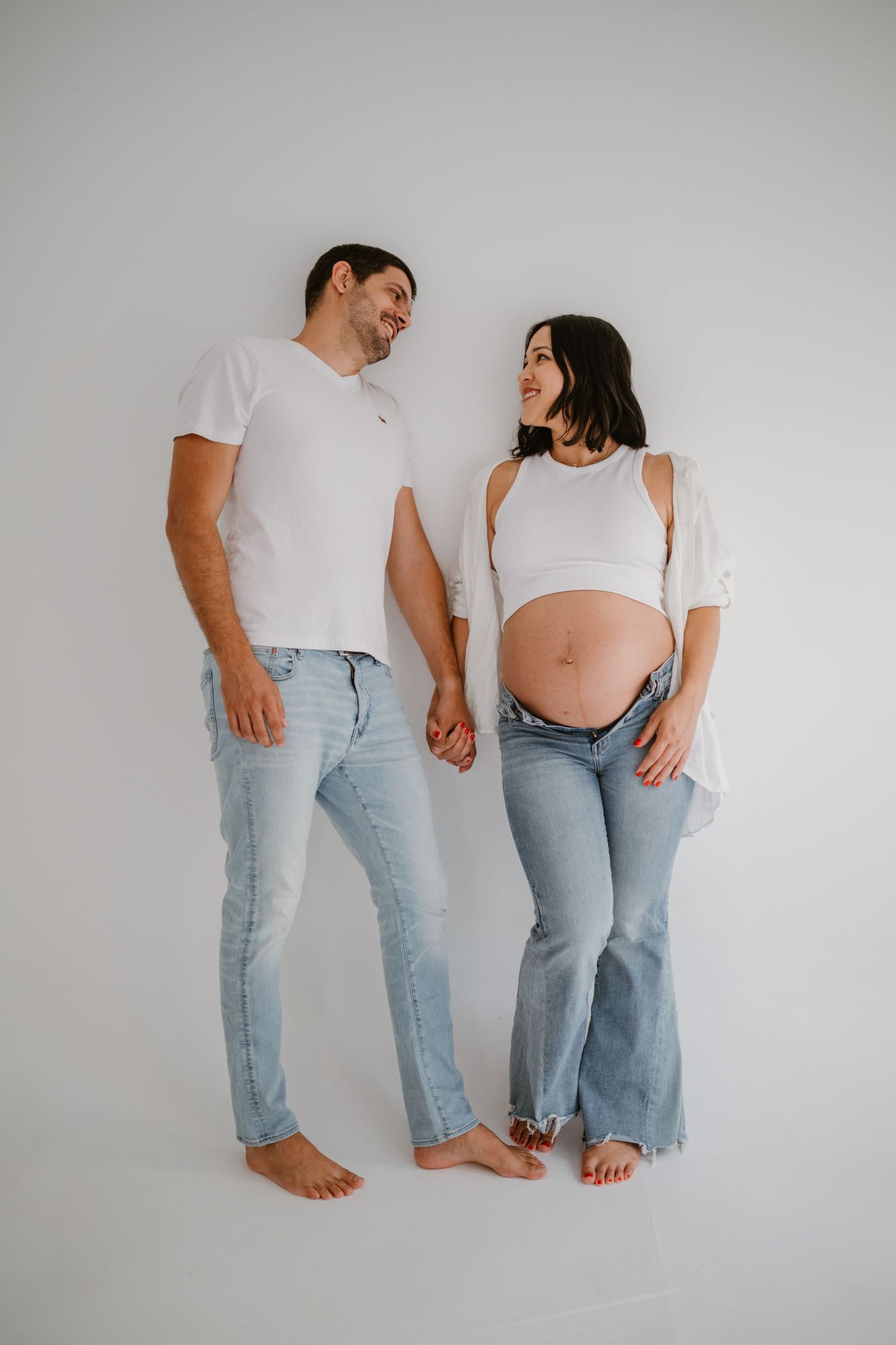Casual maternity portrait of couple in matching denim against white background.