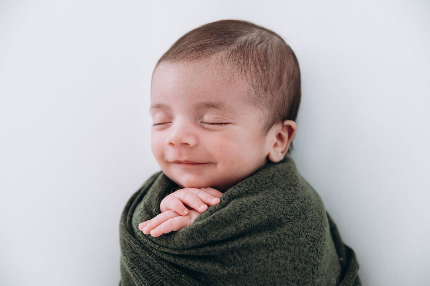 A sweet sleeping newborn baby wrapped in a dark olive green swaddle blanket against a light background.