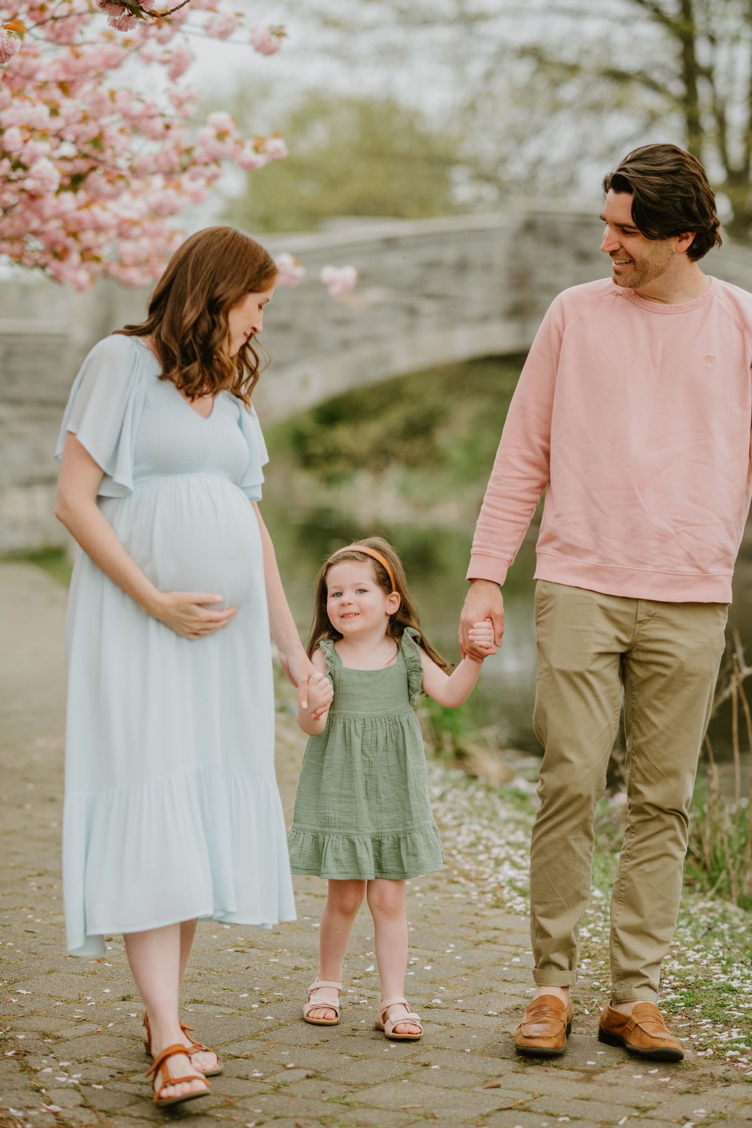 A family walking together under cherry blossoms with a pregnant mother and young daughter.