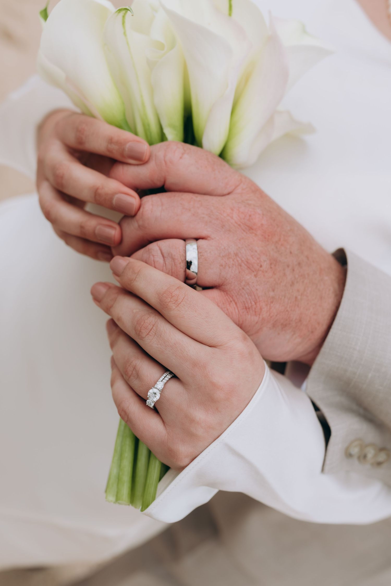 Close-up of newlyweds' hands wearing wedding rings while holding a white calla lily bouquet together.