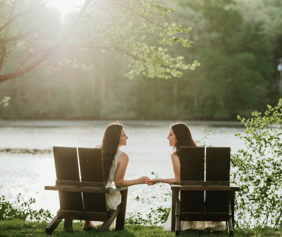 Two people sitting in Adirondack chairs by a sunlit lake having a conversation on a peaceful summer evening.