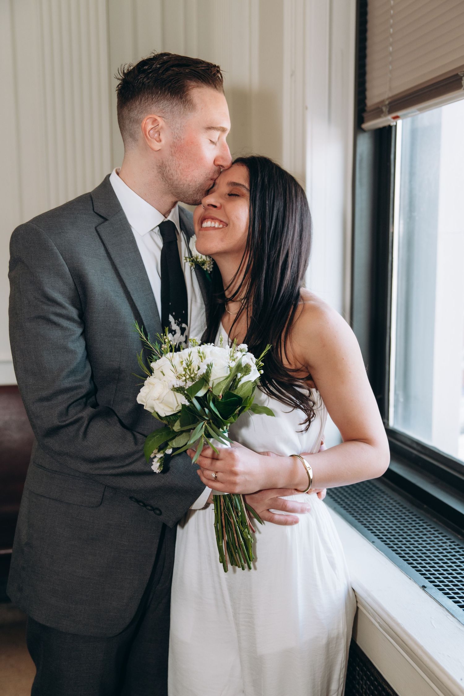 Wedding couple sharing intimate moment by window with white bouquet.