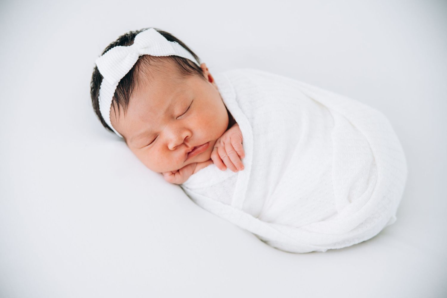 A peaceful sleeping newborn wrapped in a soft white swaddle with a striped headband rests on white bedding.