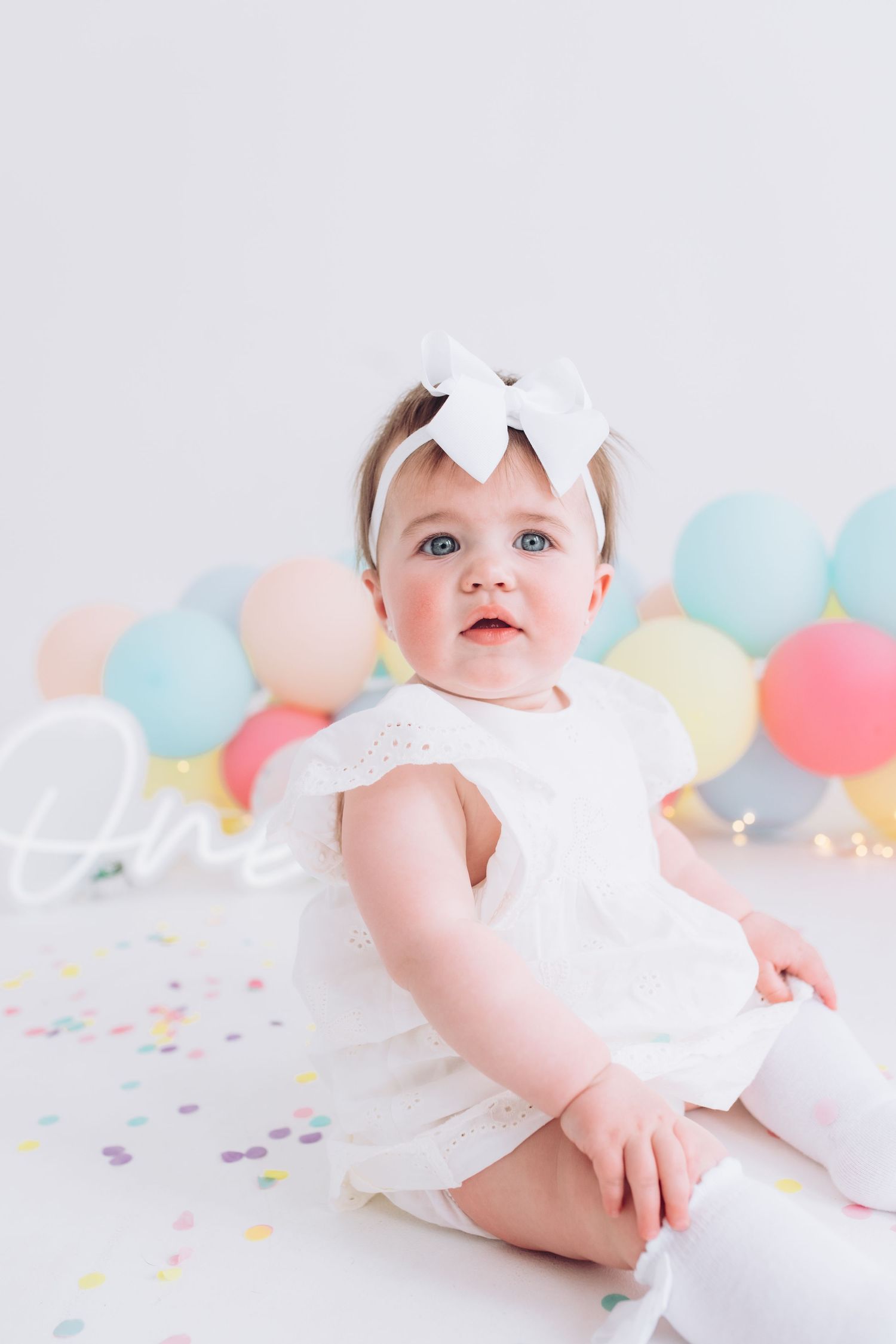 Sweet baby in white dress with bow headband sitting among pastel balloons and confetti on light background.