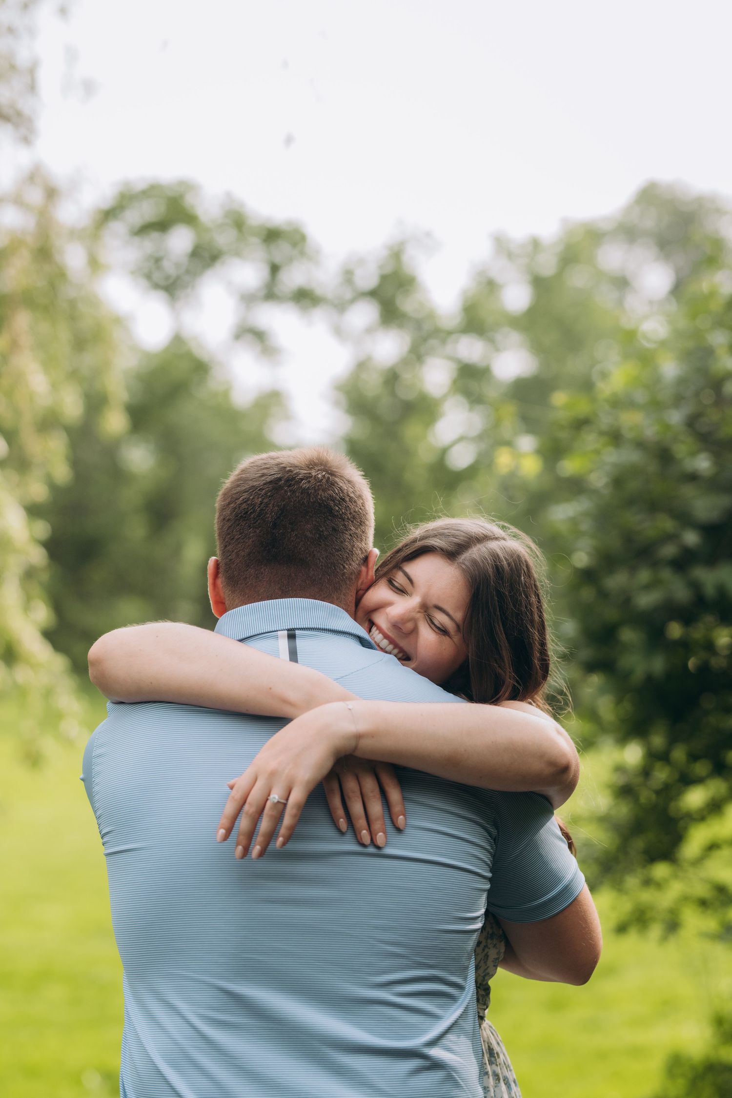 A couple embraces in an intimate hug outdoors on a sunny day with green foliage in the background.